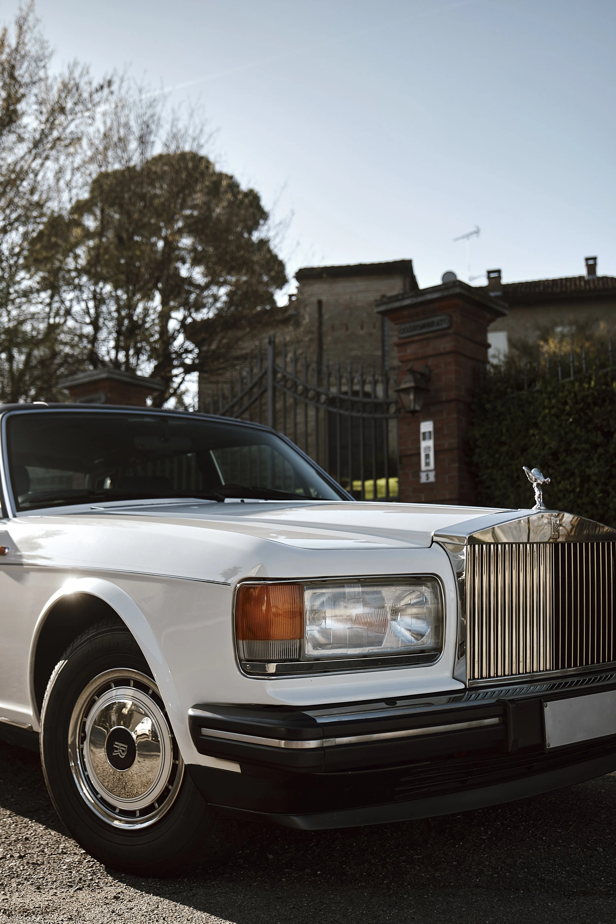 A classic white Rolls-Royce car parked outdoors near a brick gate and house, with trees in the background.