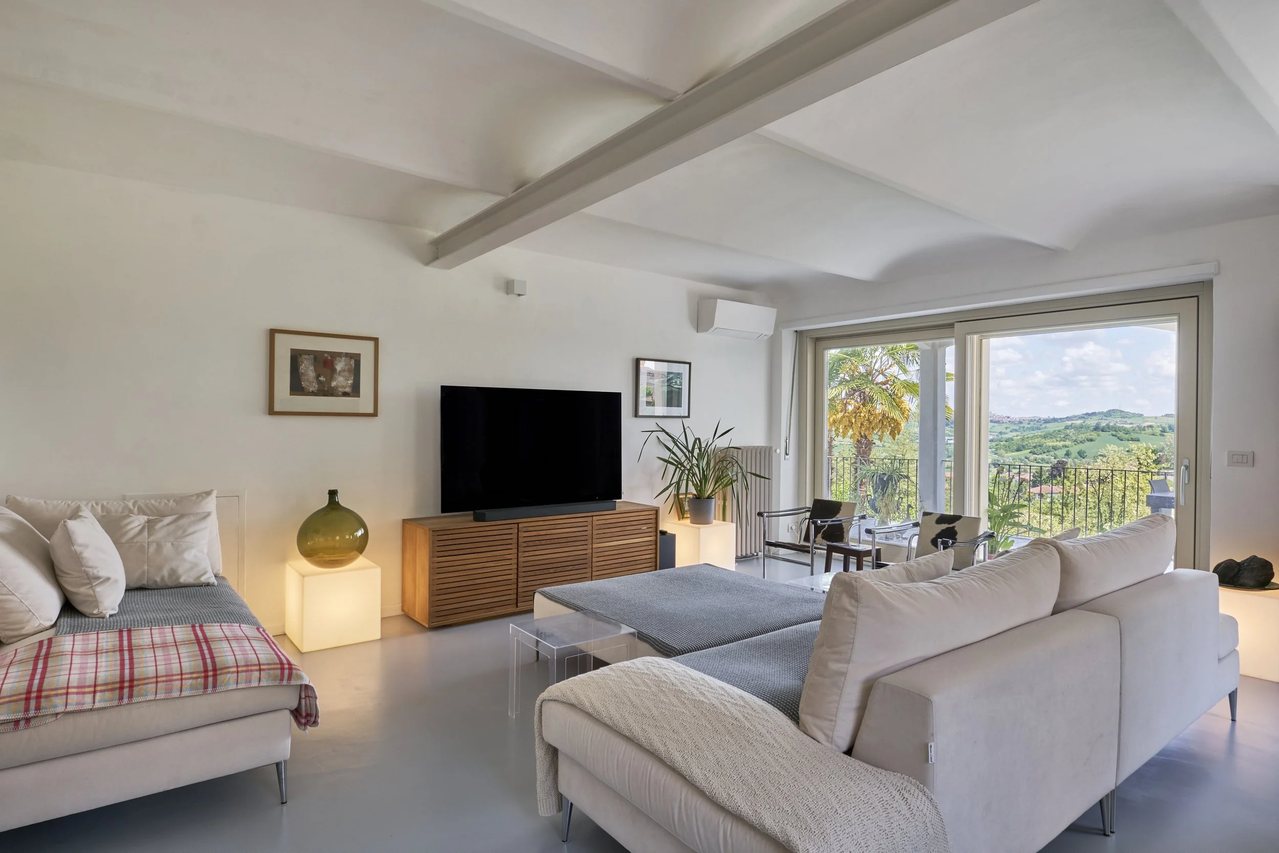 Modern living room with white walls, beige sofa, and a view of green hills through large sliding glass doors.