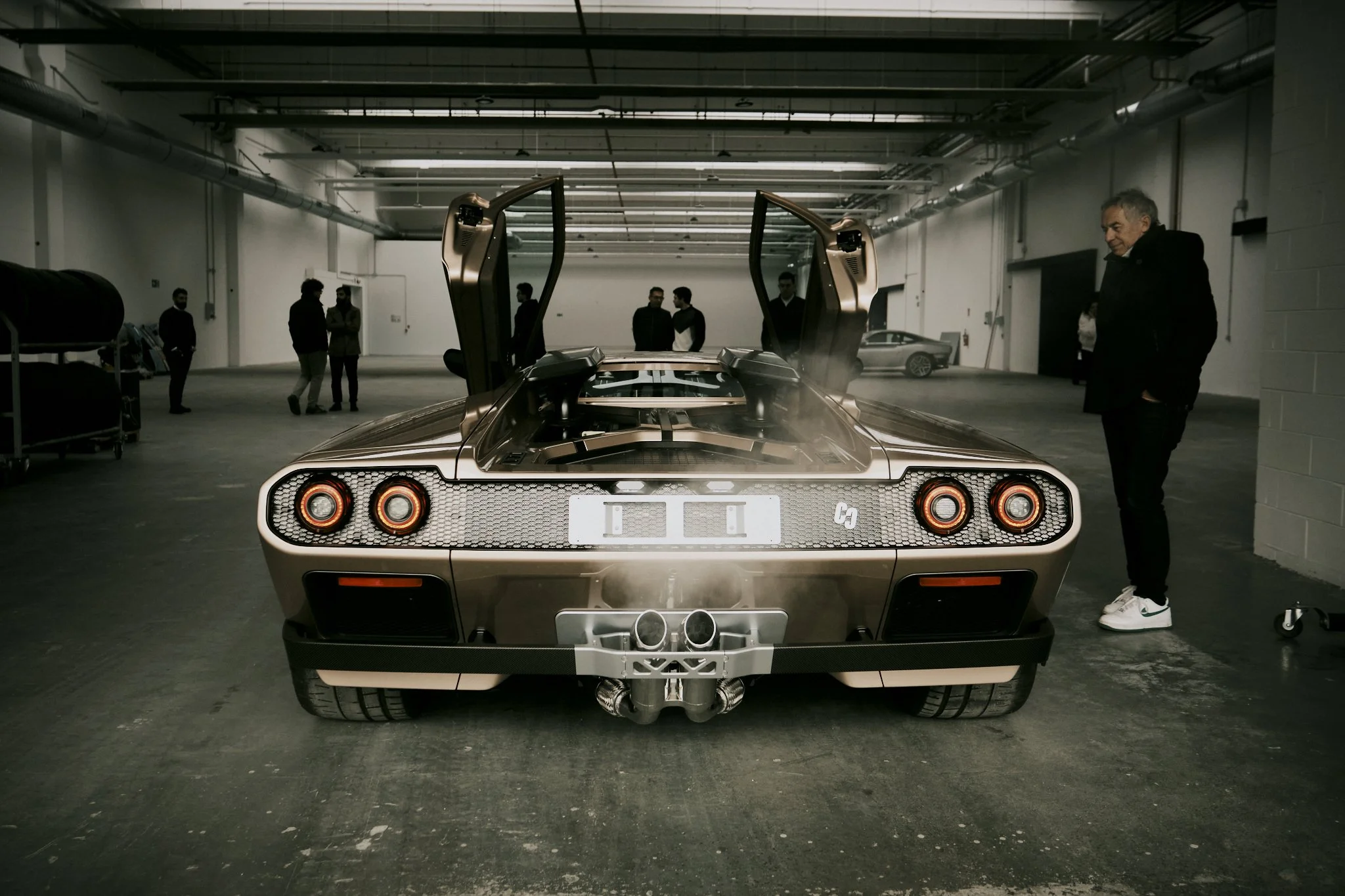 A gold-colored Lamborghini Countach with butterfly doors open in a garage, with several people observing.