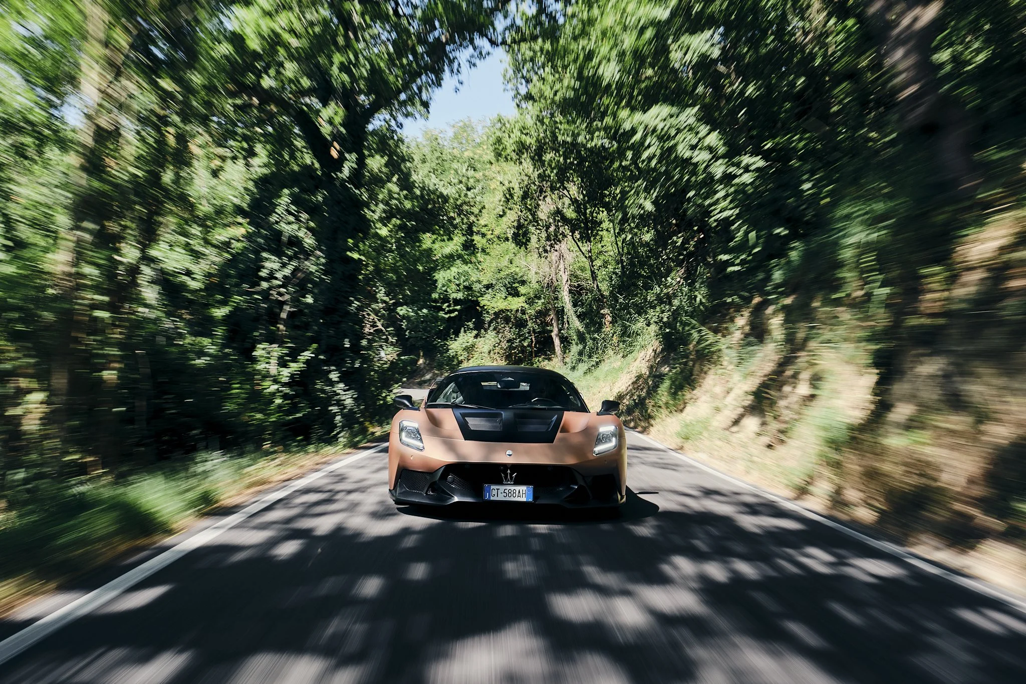 A brown sports car driving on a winding forest road with trees and greenery on both sides, and shadows from the leaves above.