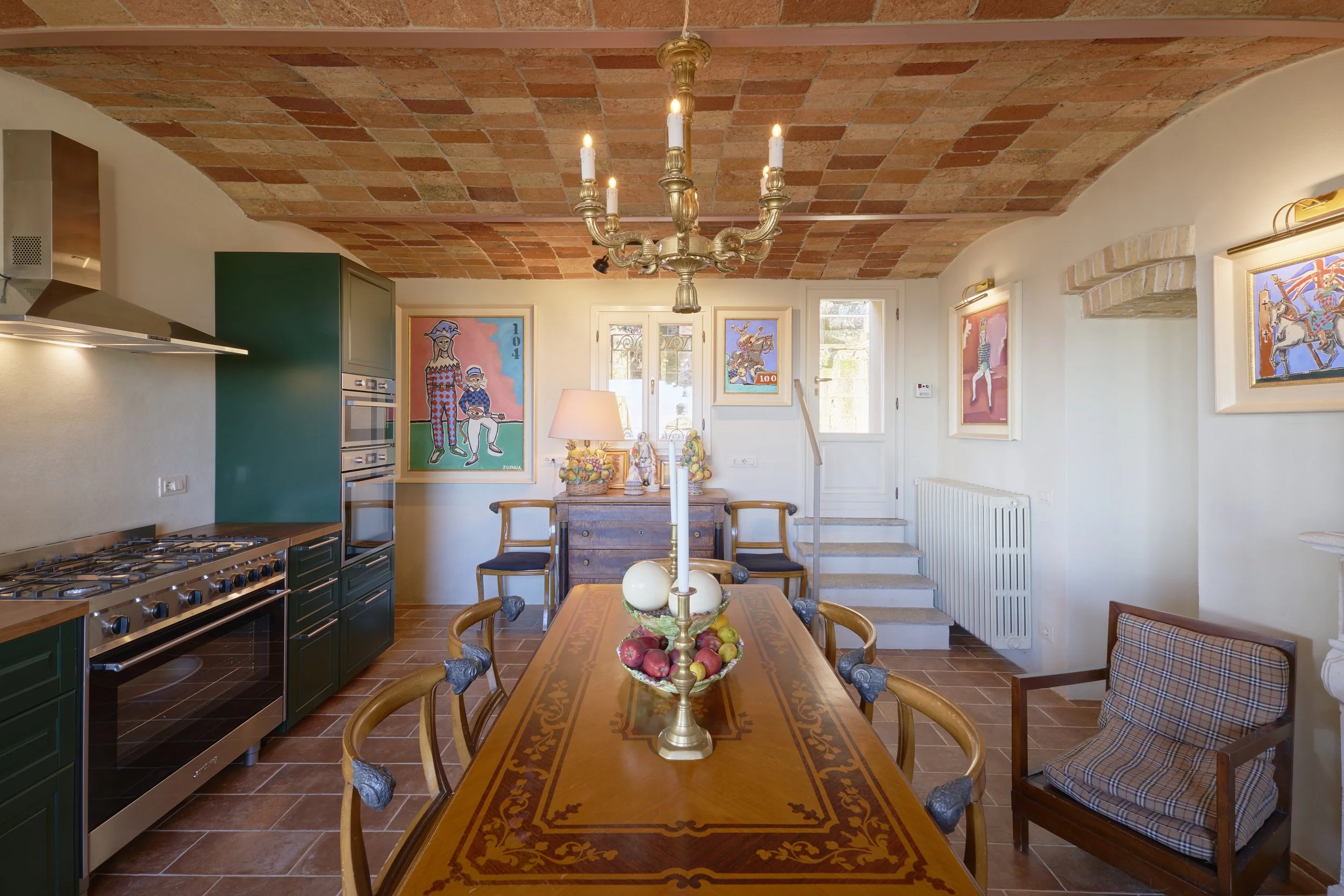 Dining room with wooden table, chairs, and a chandelier, decorated with paintings, a side table with lamps, and kitchen appliances on the left, with a staircase, window, and a radiator on the right.