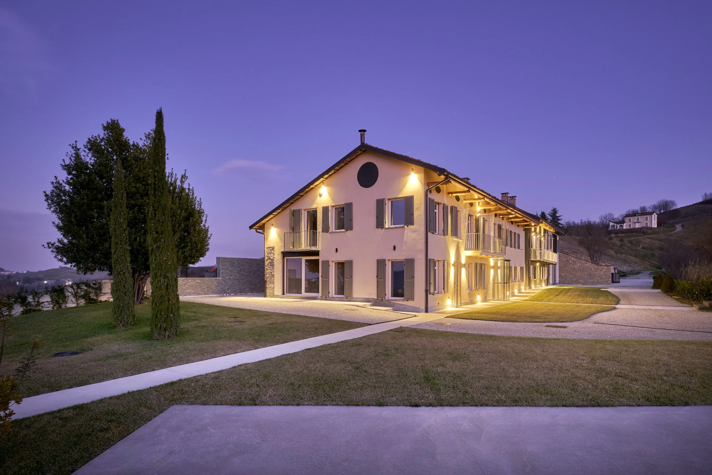 A modern two-story house with exterior lighting on a grassy area during twilight, with trees and hilly landscape in the background.