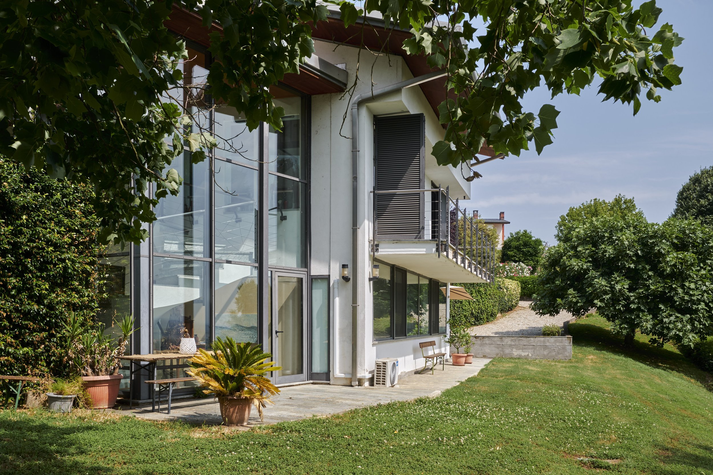 Exterior view of a modern multi-story house with large glass windows, a small balcony, and a well-maintained garden with potted plants and trees.