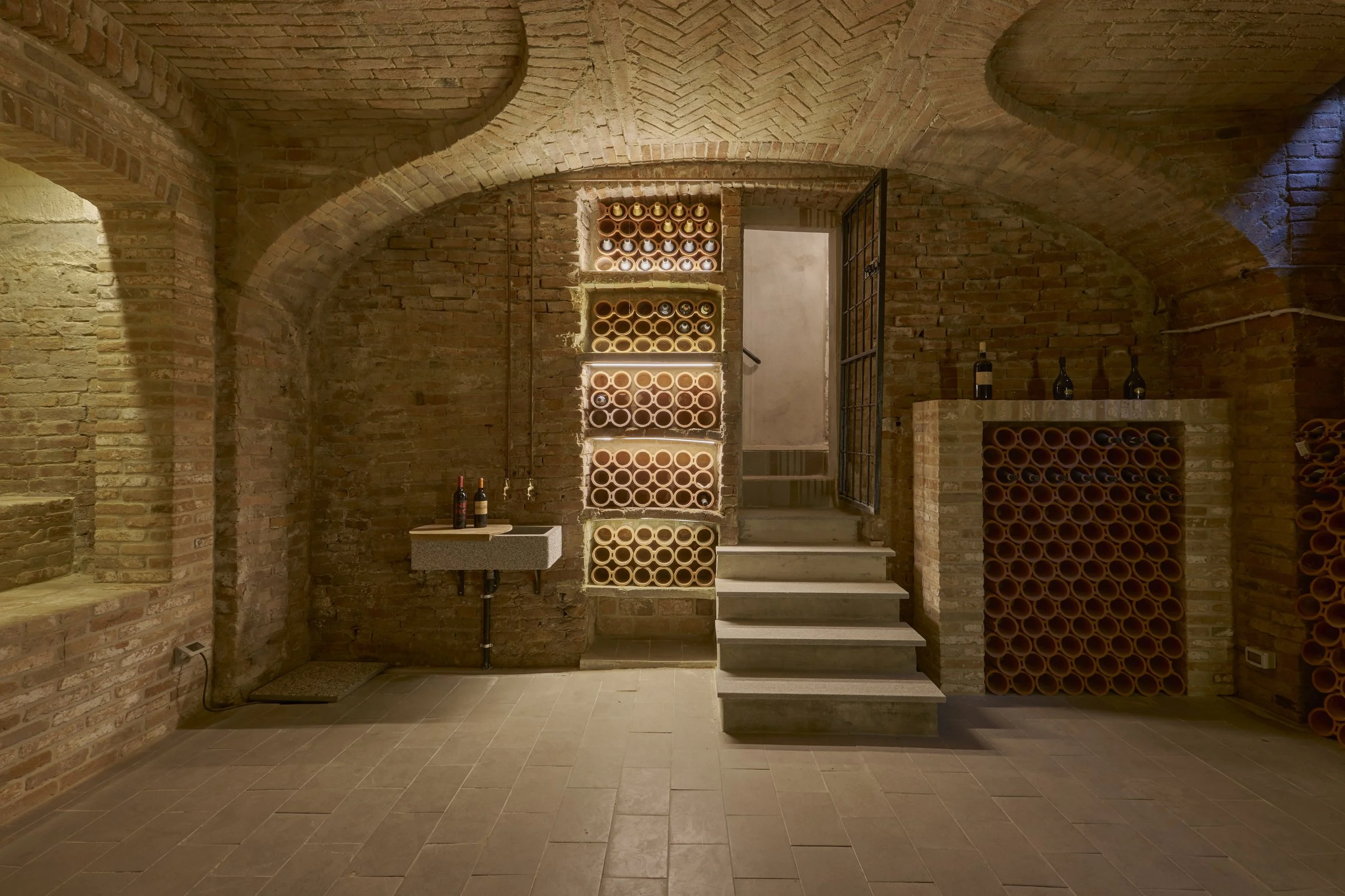 Underground wine cellar with brick walls, arched brick ceilings, and built-in wine racks filled with wine bottles, illuminated by warm lighting.