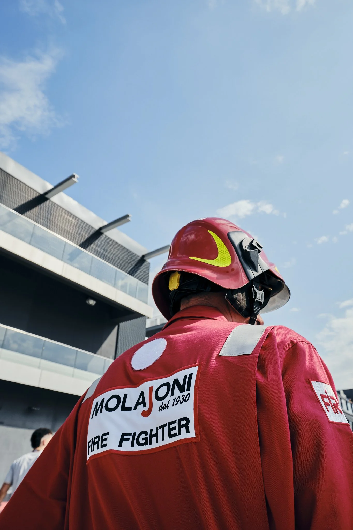 Firefighter wearing a red helmet and uniform with 'MOLAJONI' patch, standing outdoors against a blue sky and modern building.