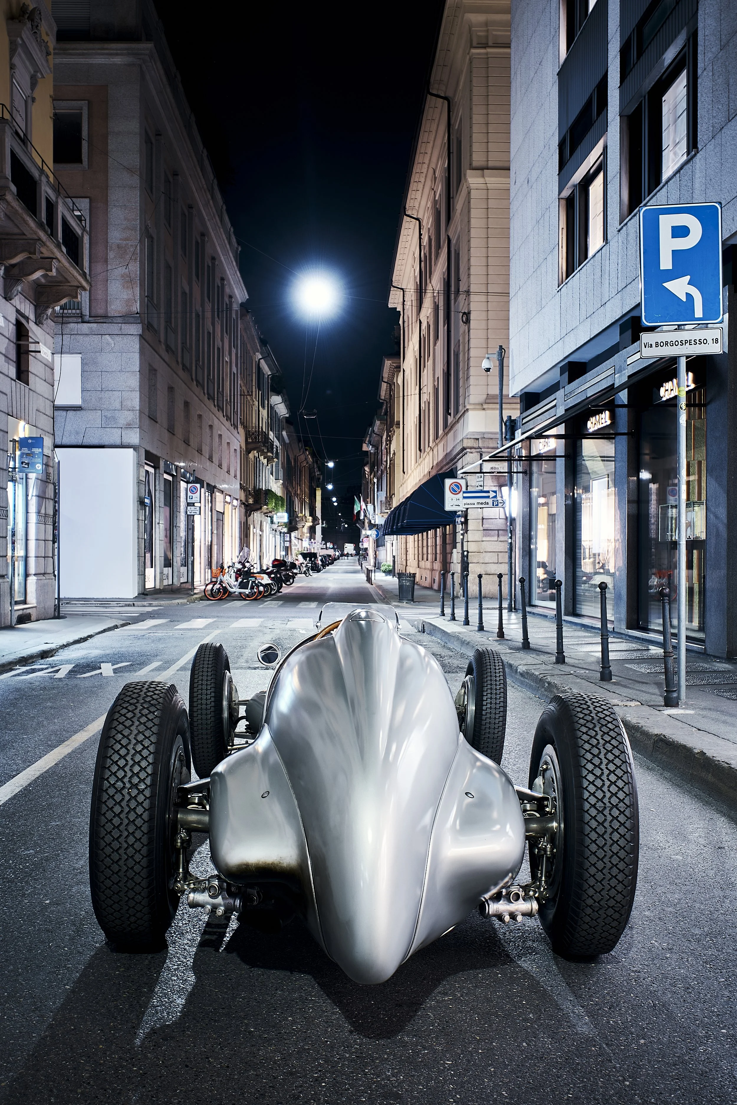 A sleek, vintage silver race car parked on a nighttime city street with illuminated buildings and a bright moon overhead.