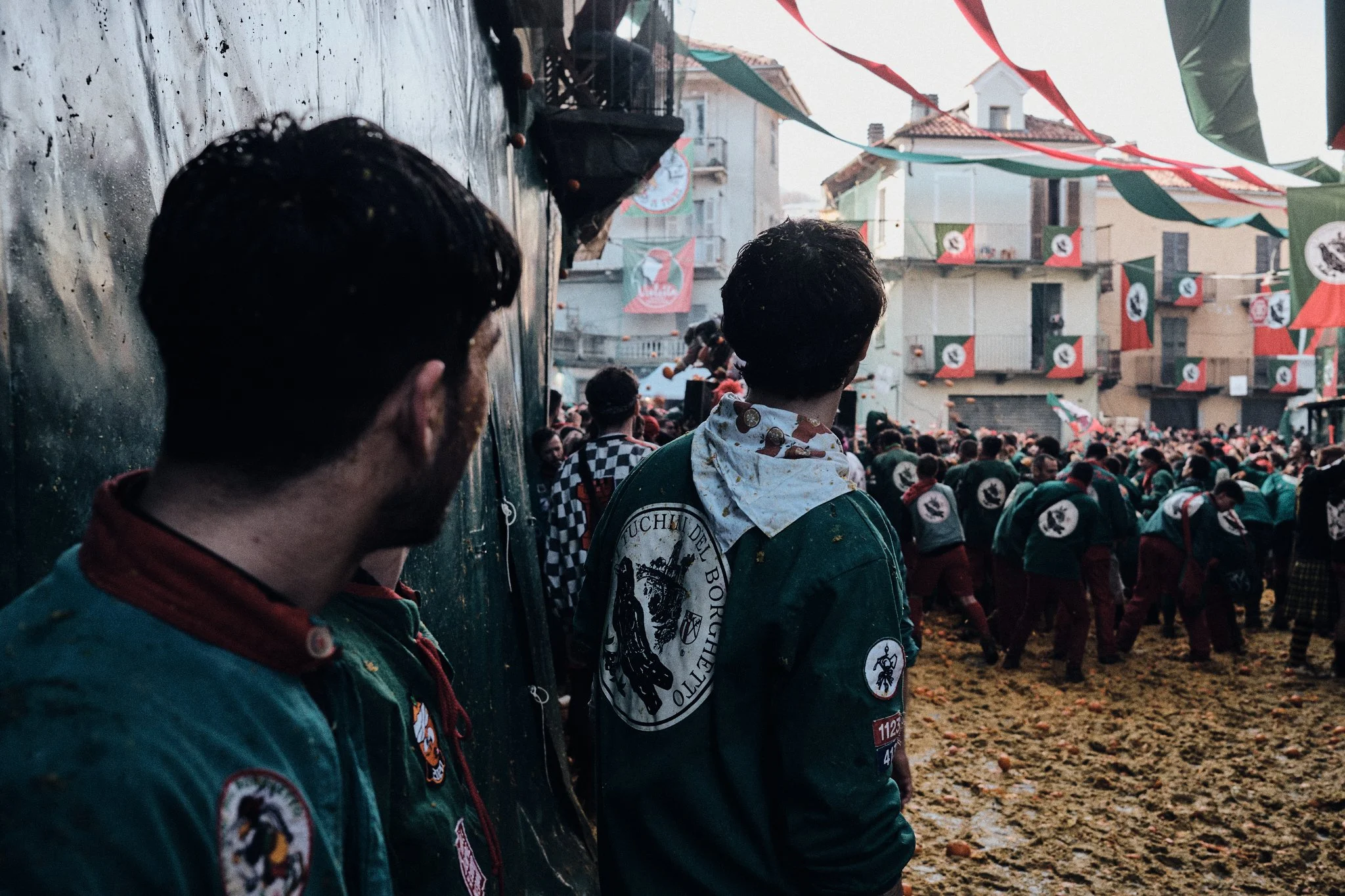 People participating in a traditional festival with flags and banners, some wearing uniforms with patches, in a crowded outdoor street scene.