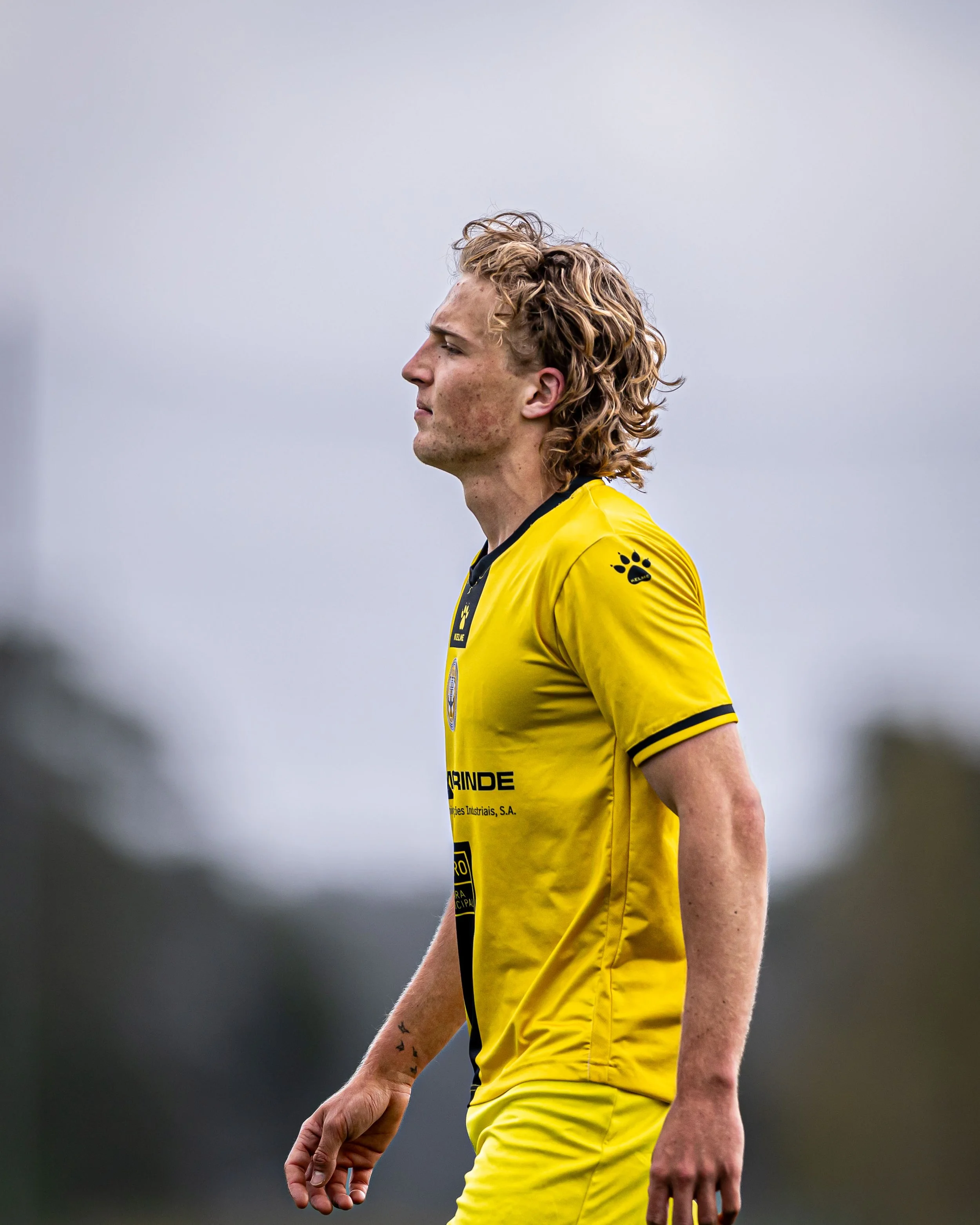 A young male soccer player with curly blonde hair wearing a yellow sports jersey with black accents, standing on a field with a cloudy sky in the background.
