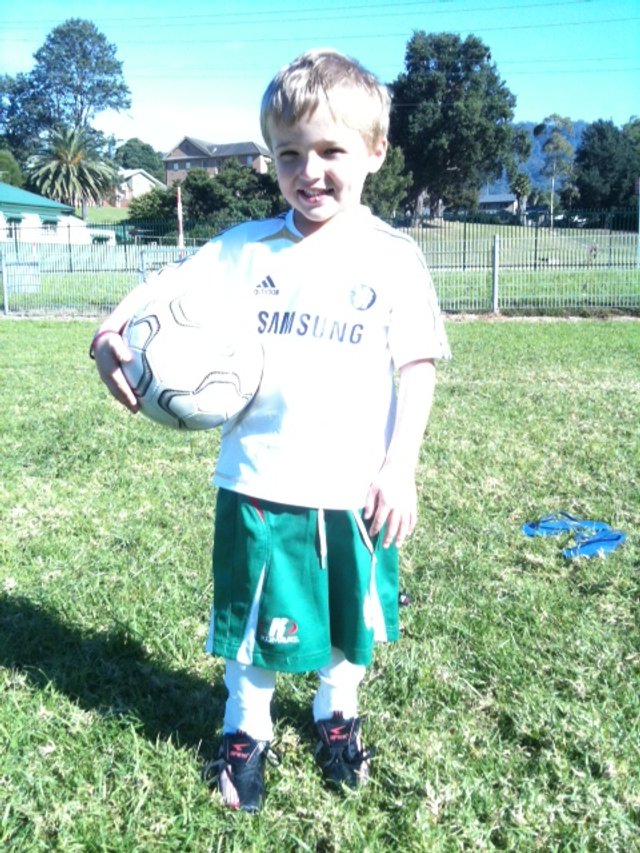 A young boy holding a soccer ball, standing on a grassy field in a park with trees and houses in the background.
