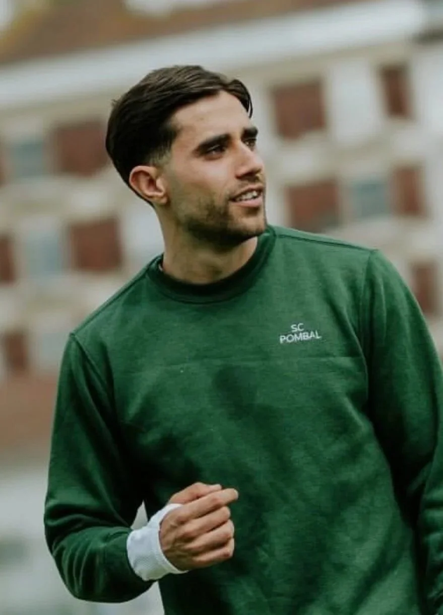A young man with dark hair and beard wearing a green sweatshirt with 'SC POMBEAL' on it, standing outdoors near a building.