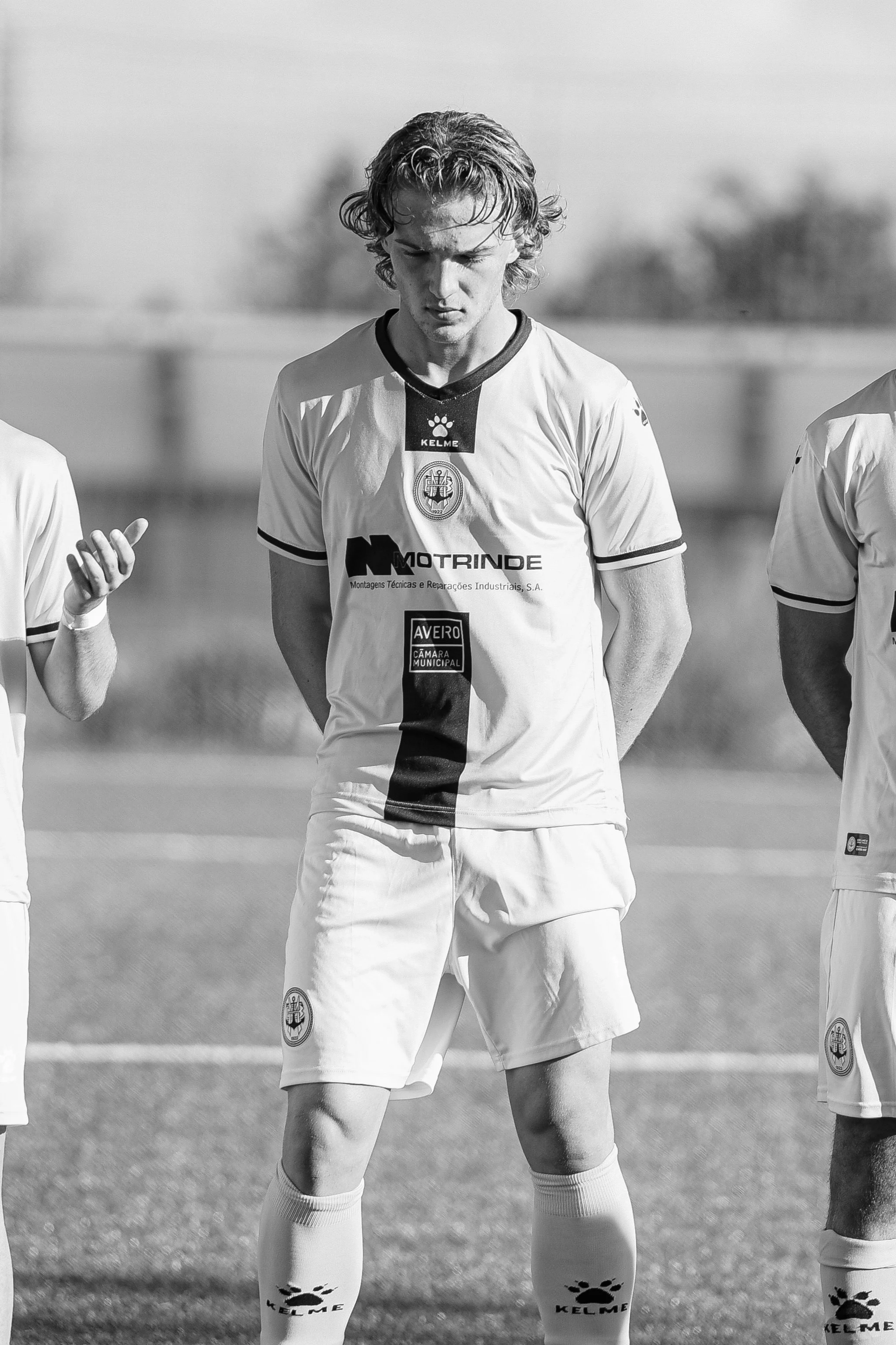 Black and white photo of a male soccer player in a uniform, standing with hands behind his back, on a soccer field.
