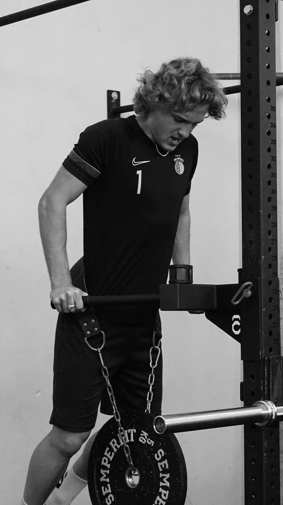 A person with curly hair in a black sports jersey is in a gym, lifting a barbell with a chain and weight plate while attached to a power rack.