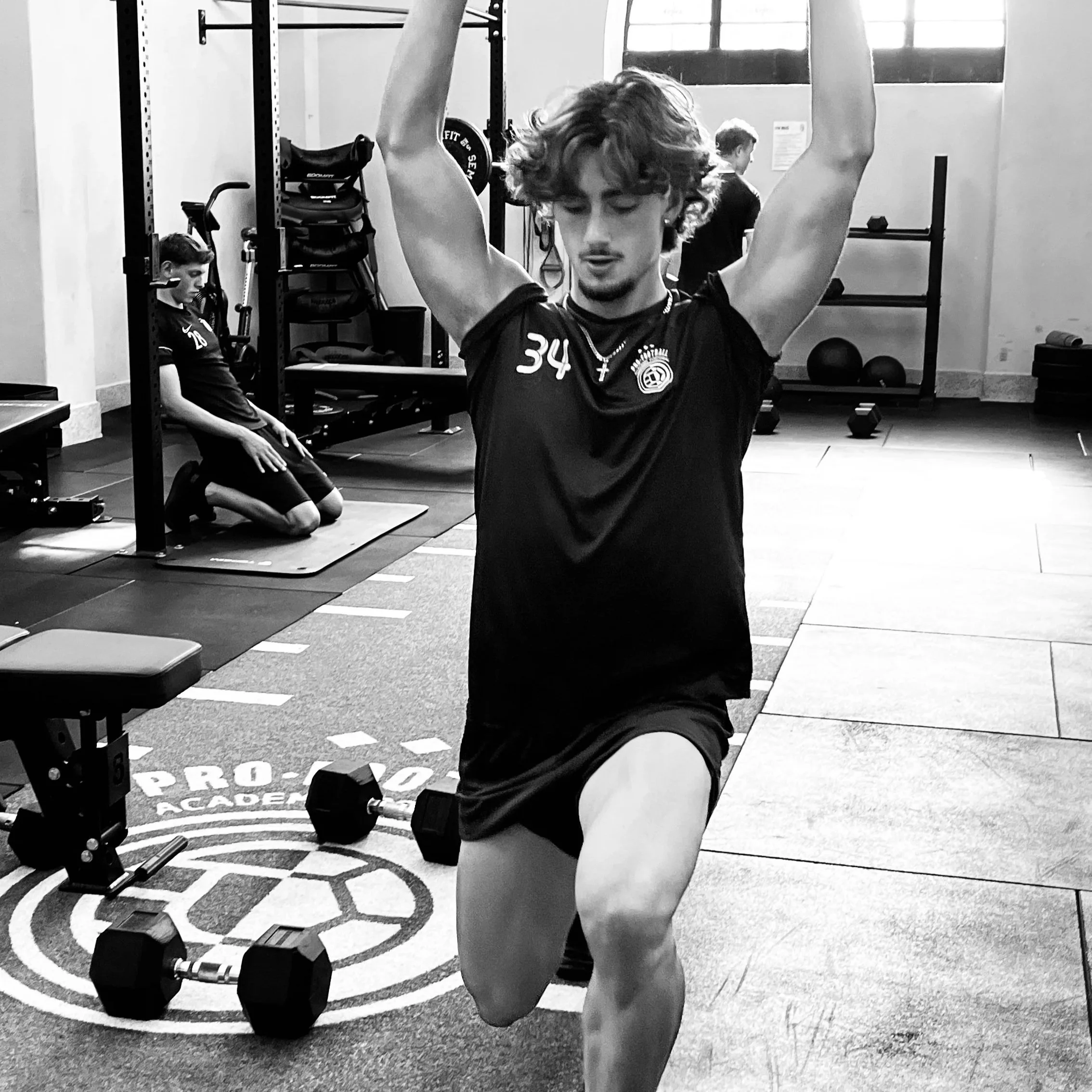 Young man performing a lunge exercise with arms raised in a gym, with weights and people in the background.