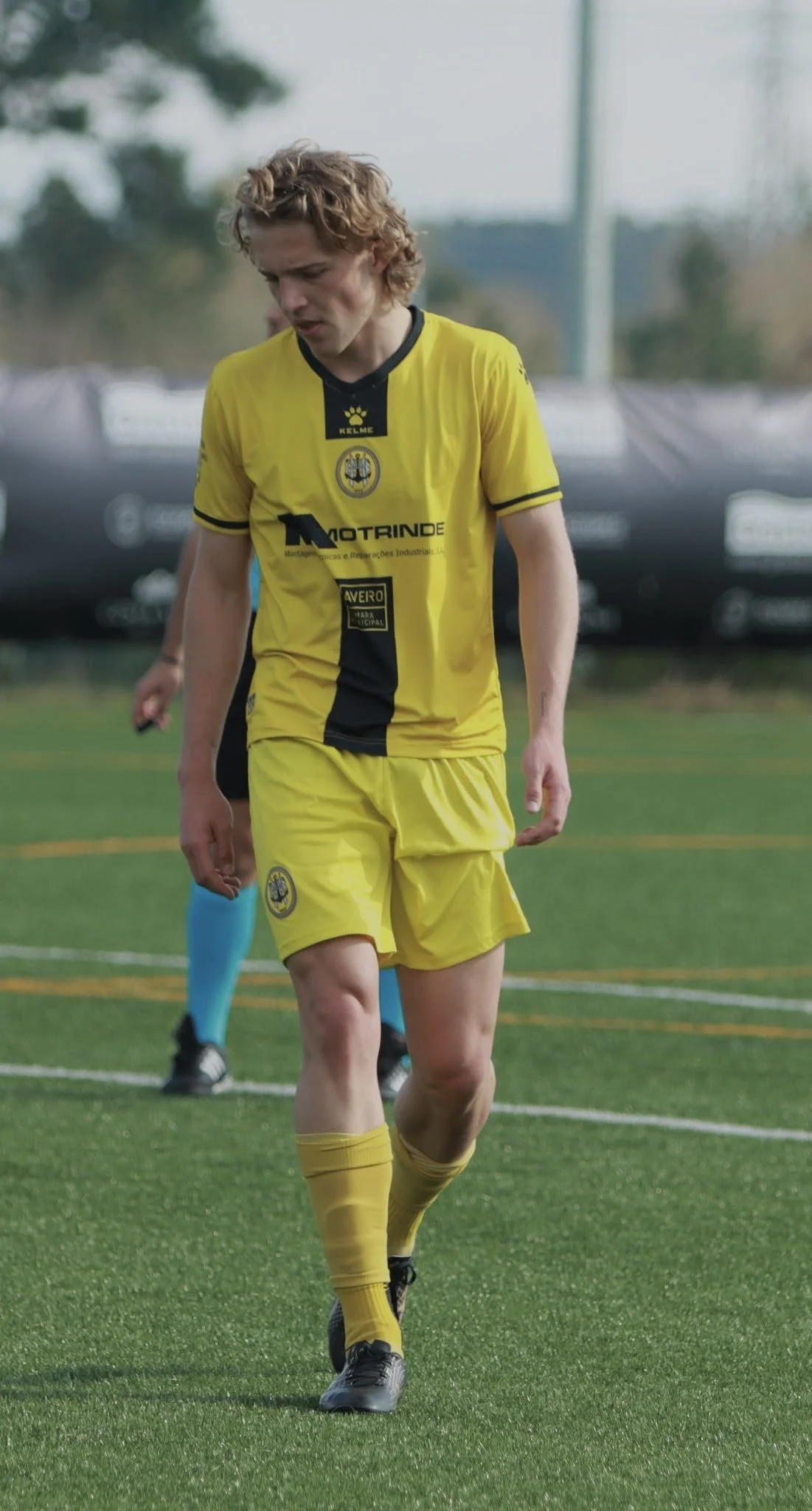 A soccer player in a yellow uniform standing on a green field, looking down with a contemplative expression.