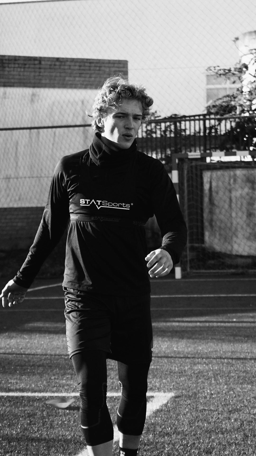 Black and white photo of a young male athlete running on a sports field, wearing athletic clothing including a long-sleeve top and leggings.