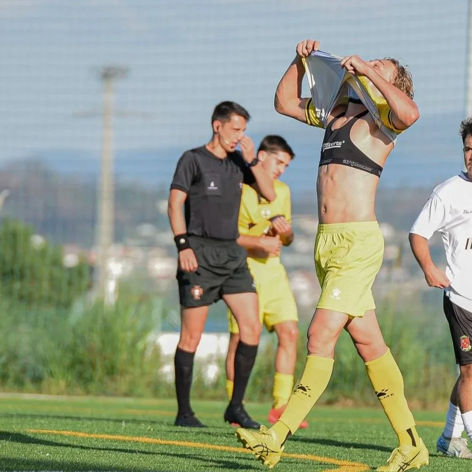 A soccer player in yellow shorts and shirt removing his jersey on the field, with two other players and a referee in the background.