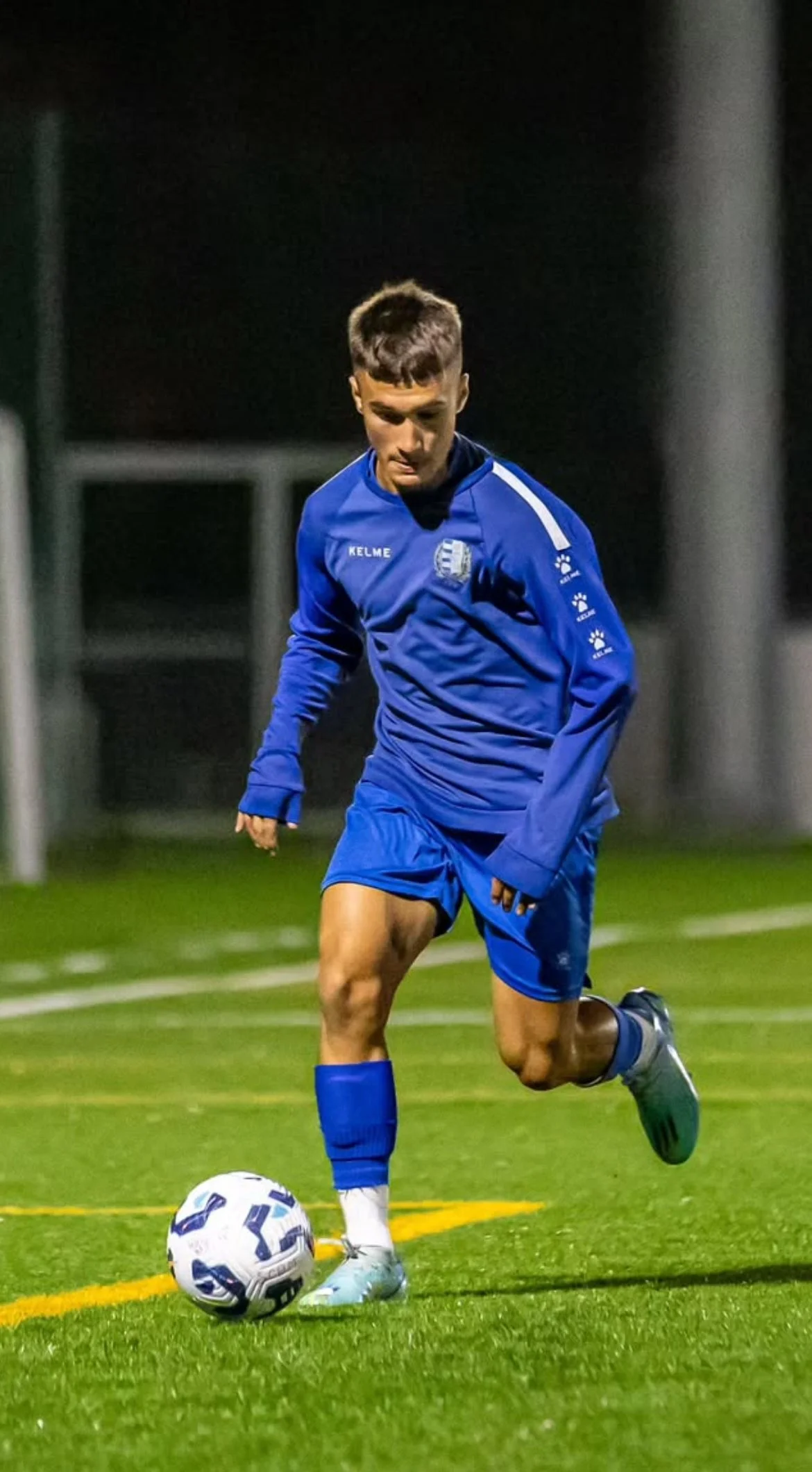 A young male soccer player in a blue uniform practicing on a green turf field at night, about to kick a soccer ball.