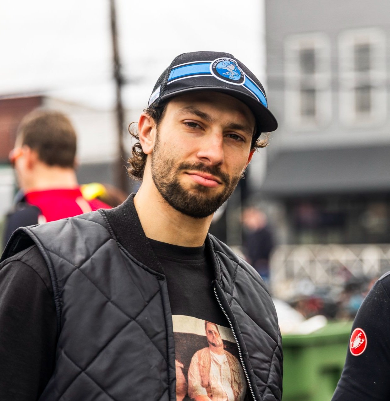 A man with a beard and mustache wearing a black quilted jacket and a black baseball cap with a blue emblem stands outdoors in an urban setting.