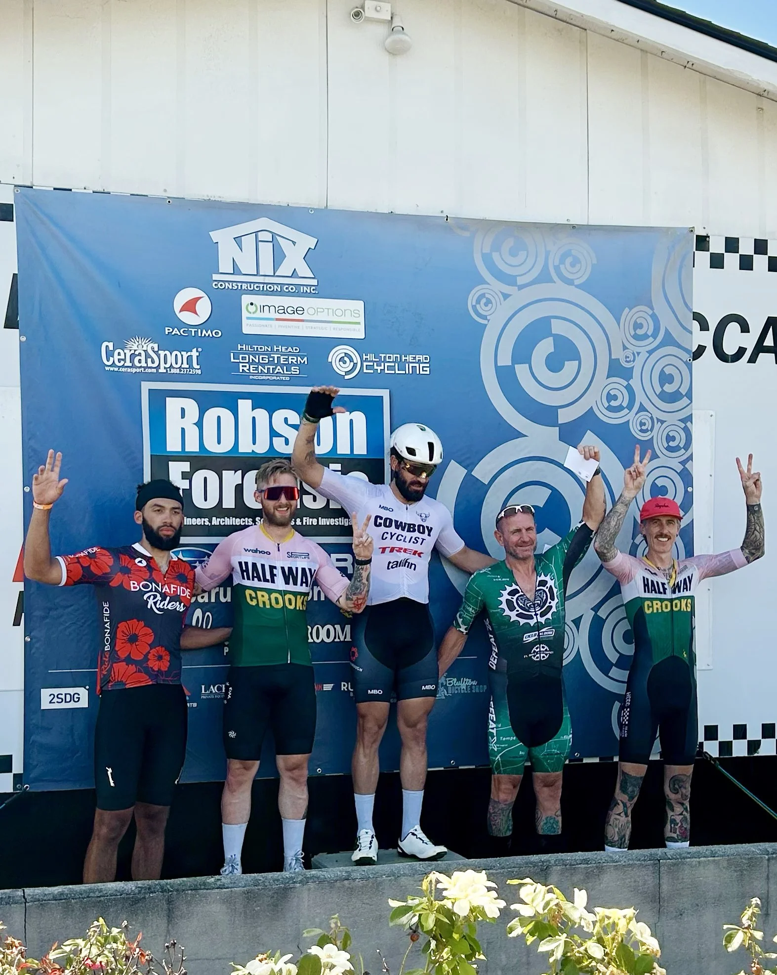 Cyclists on a podium celebrating after a race, with five men holding up peace signs and wearing cycling jerseys and gear, standing in front of a blue event backdrop.