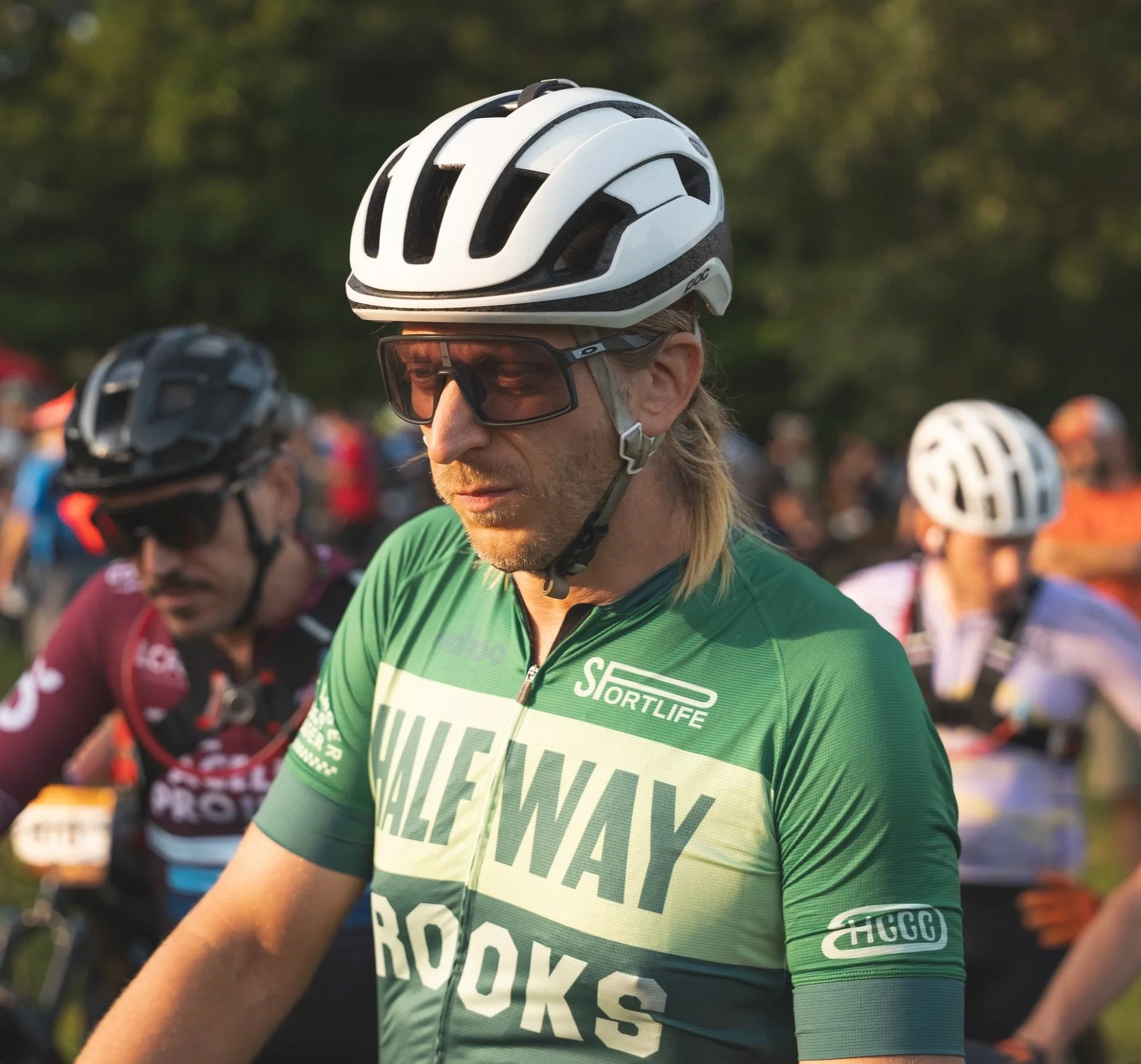 Cyclist wearing a green jersey with 'Halfway Crooks' and logo, white helmet, and sunglasses, preparing for a race along with other cyclists in the background.