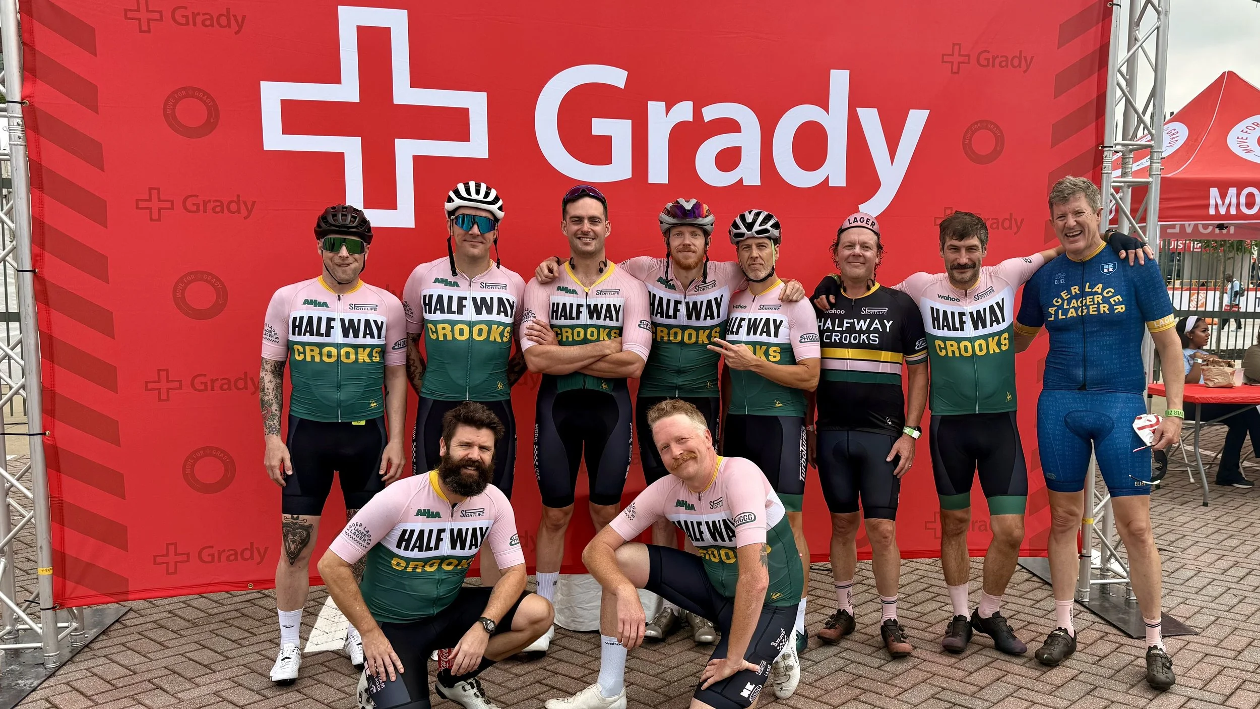 Group of cyclists in matching jerseys and helmets, standing in front of a red backdrop with the "Grady" logo, celebrating after a cycling event.