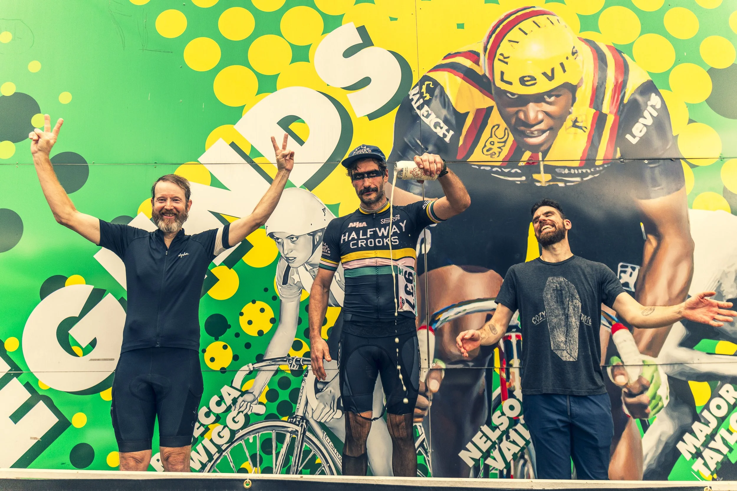 Three men celebrating on stage with a large colorful background featuring cycling imagery. The man in the middle, dressed in cycling gear, drinks from a bottle, while the men on either side cheer with raised arms.