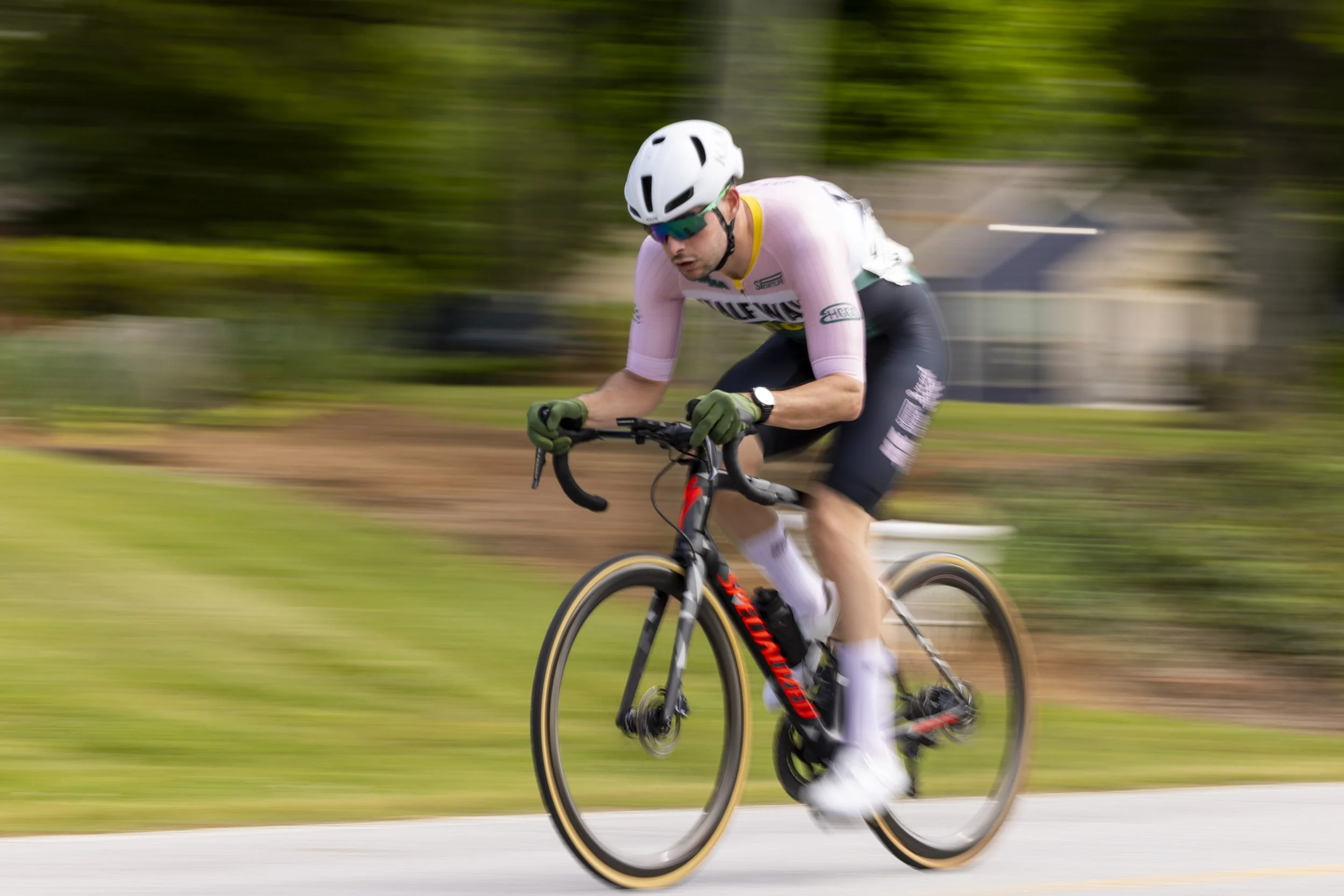 A cyclist wearing a white helmet, sunglasses, and a pink and black cycling outfit rides a black and red bike on a road with a blurred green background.