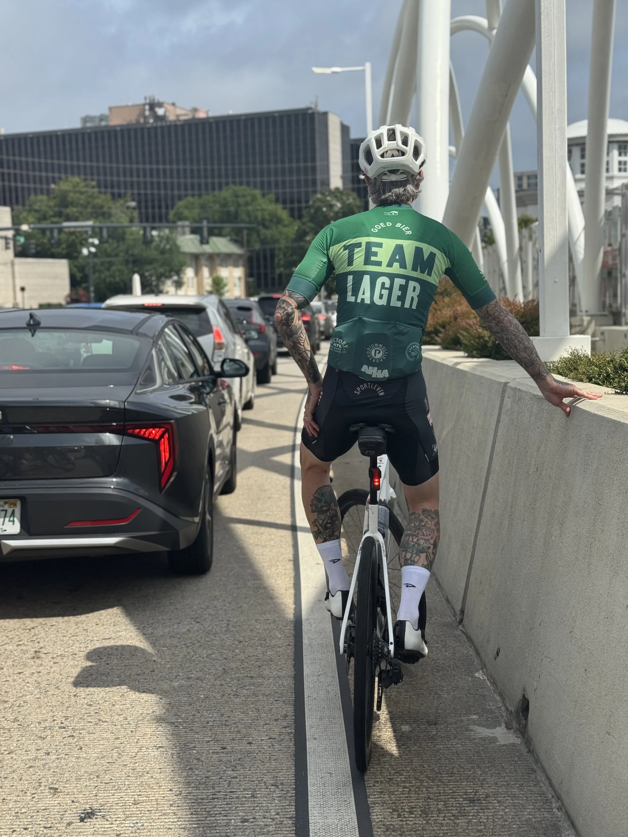 A person with tattoos wearing a green cycling jersey and helmet, riding a white bicycle next to a concrete barrier on a city street, with cars and modern buildings in the background.