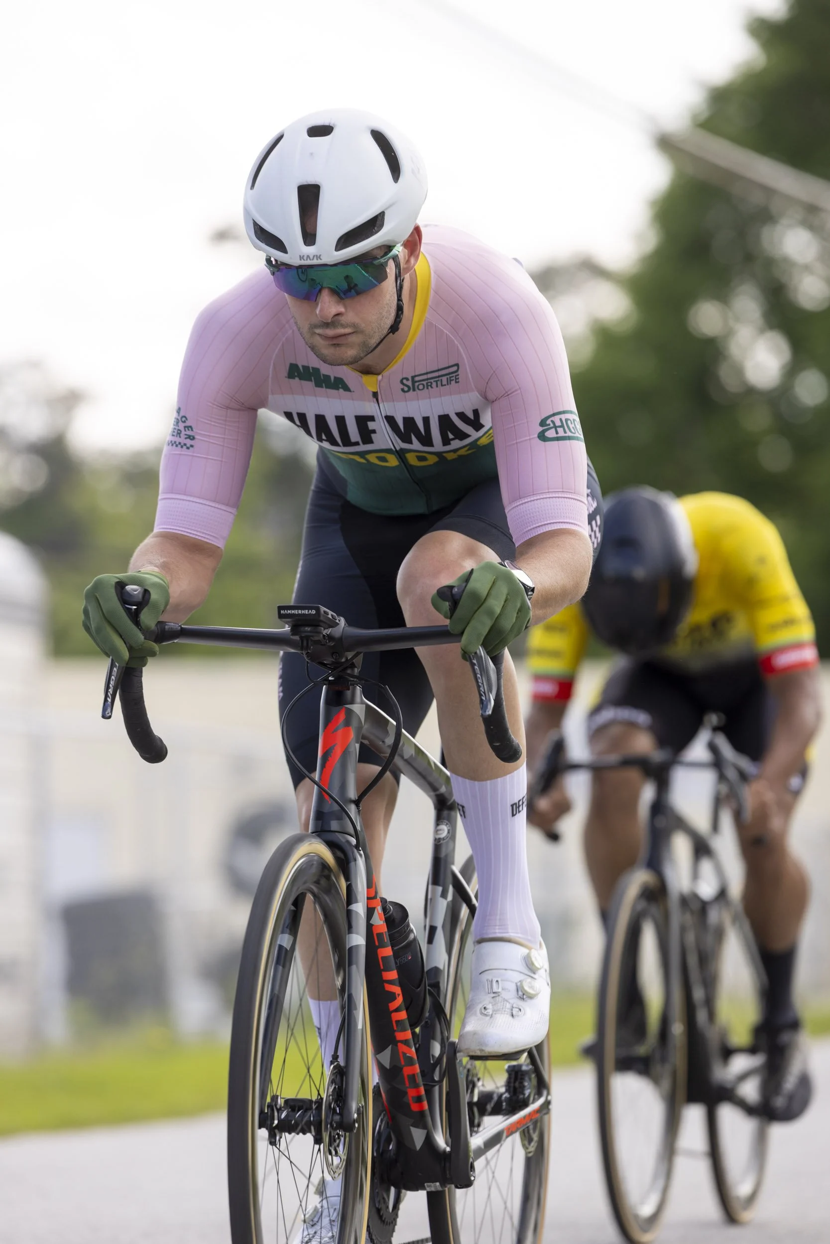 Cyclist wearing white helmet, sunglasses, green gloves, pink and green jersey, and white shoes riding a black and red Specialized bicycle, with another cyclist in yellow and black behind him.