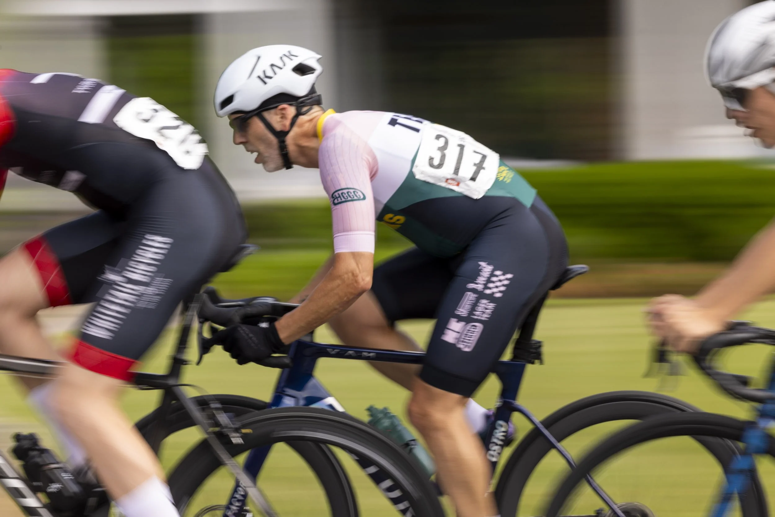 Cyclists racing in a competition with a blurred background, focused on a man wearing a white helmet, pink and green jersey, and black shorts with the number 317.