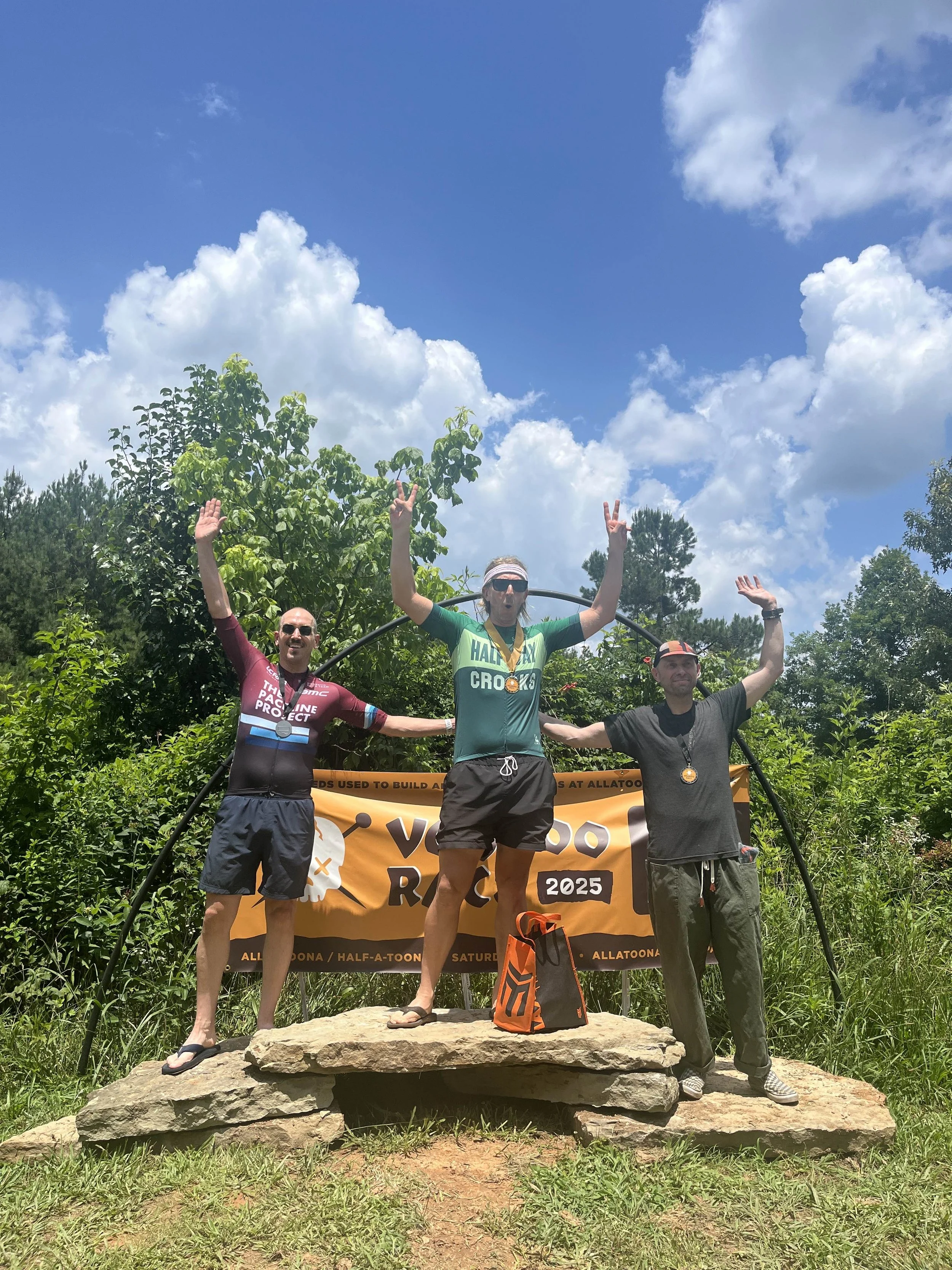 Three men standing on a rock pedestal celebrating after a race, with one in the center (on the top of the podium wearing HALFWAY CROOKS kit) and two on either side, in front of a banner that says 'VOODOO RACE 2025'. 