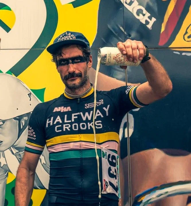 A man in cycling gear with face paint, holding a can of drink, standing in front of a colorful background with large graphics.