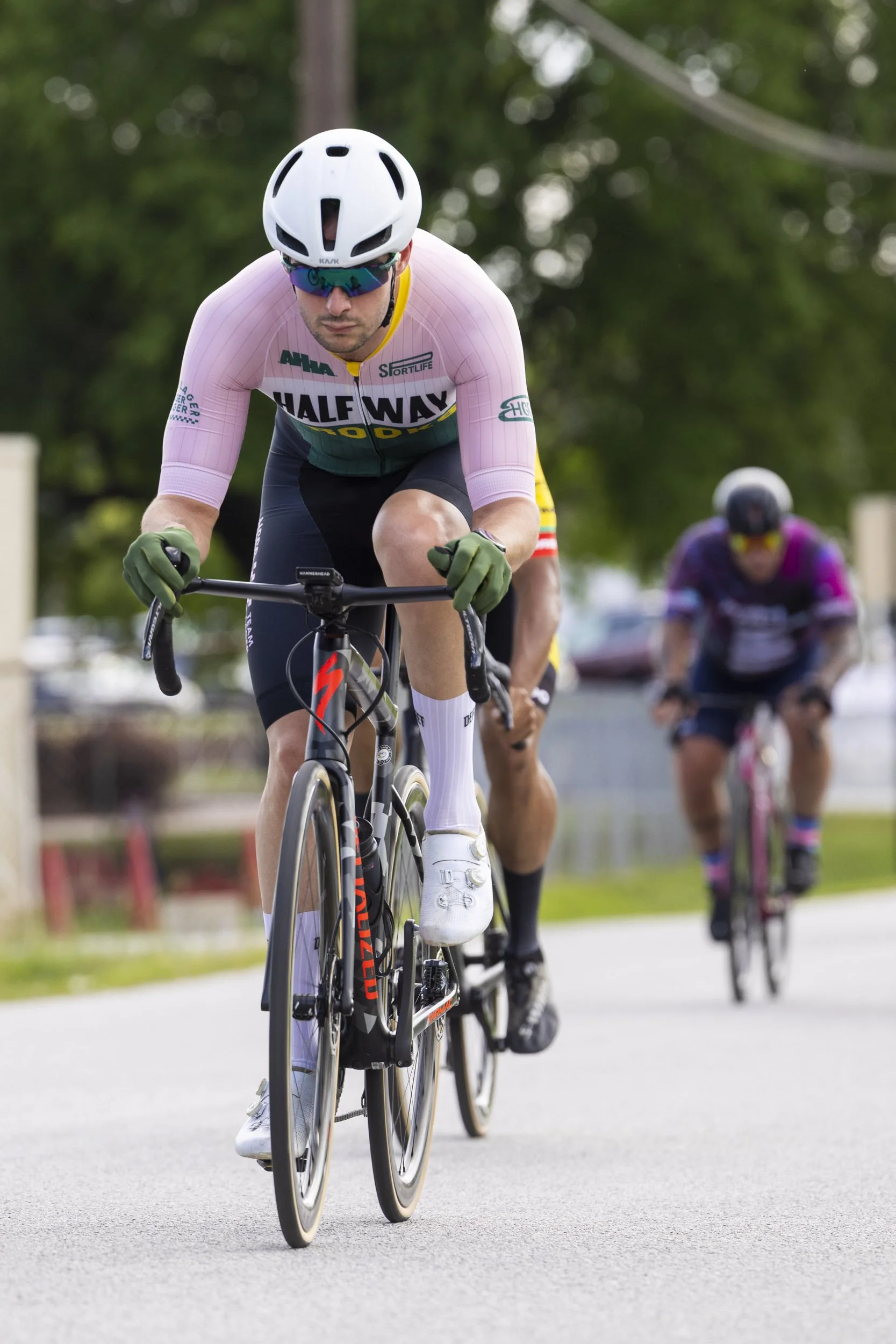 A cyclist wearing a white helmet, sunglasses, a pink and green cycling jersey, and black shorts is riding a black road bike with a red logo. There are two other cyclists behind him, all riding on a paved path with trees and vehicles in the background