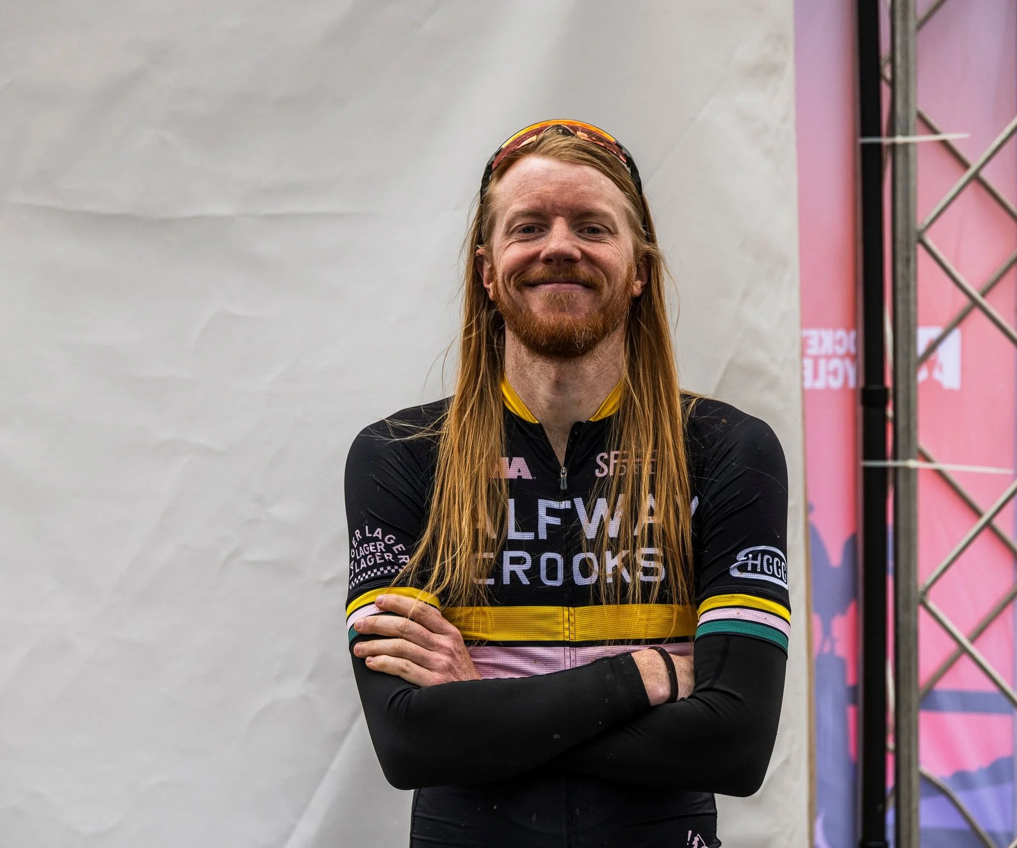 A male cyclist with long red hair and a beard, smiling, wearing a black cycling jersey with various sponsor logos, standing with arms crossed in front of a plain background.