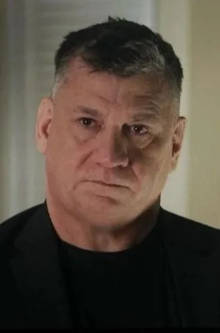 A middle-aged man with short dark hair, wearing a black shirt and a black jacket, looking directly at the camera with a serious expression.