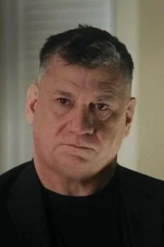 A middle-aged man with short dark hair, wearing a black shirt, looking serious or stern.
