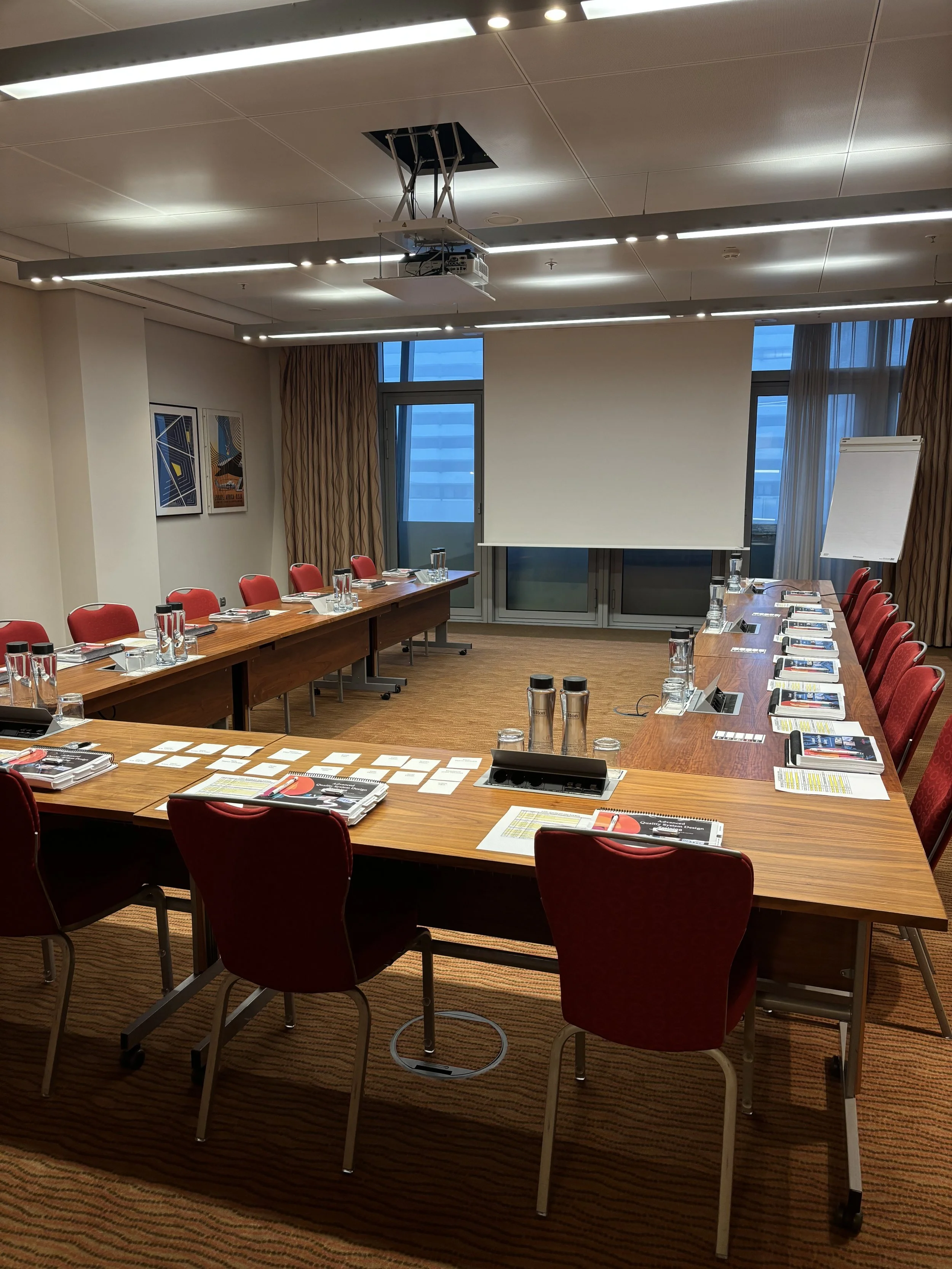 A conference room with a rectangular wooden table surrounded by red chairs, set with water glasses, notepads, and pens. A white projection screen and a flip chart are at one end of the room. There are large windows with curtains, and a ceiling-mounted projector is visible.