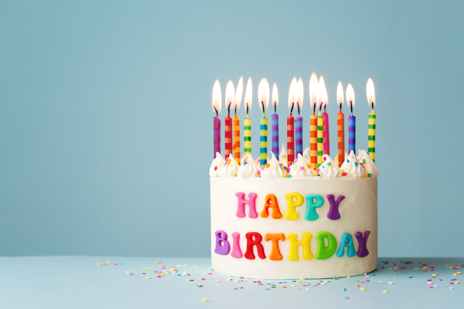 Colorful birthday cake with rainbow-striped candles and 'HAPPY BIRTHDAY' decoration, on a light blue background with scattered confetti.