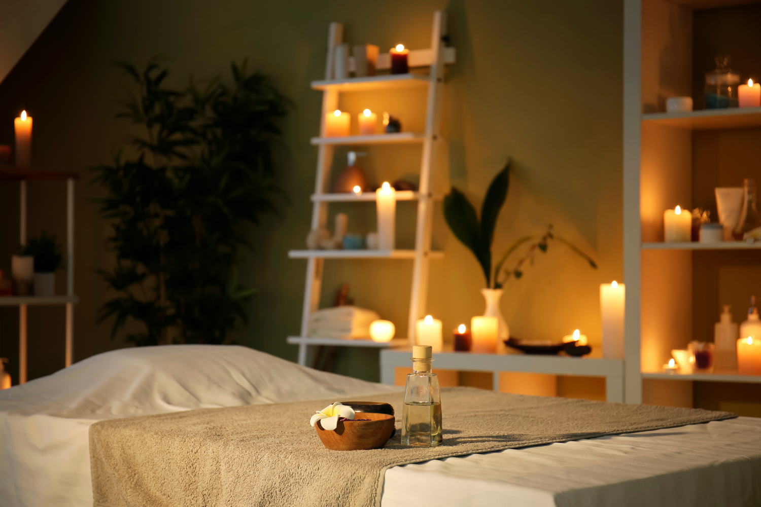 Spa or massage room with a bed covered in white linens, surrounded by lit candles on shelves and a small table, with a bottle of oil, a bowl with white flowers, and a black brush on the bed.
