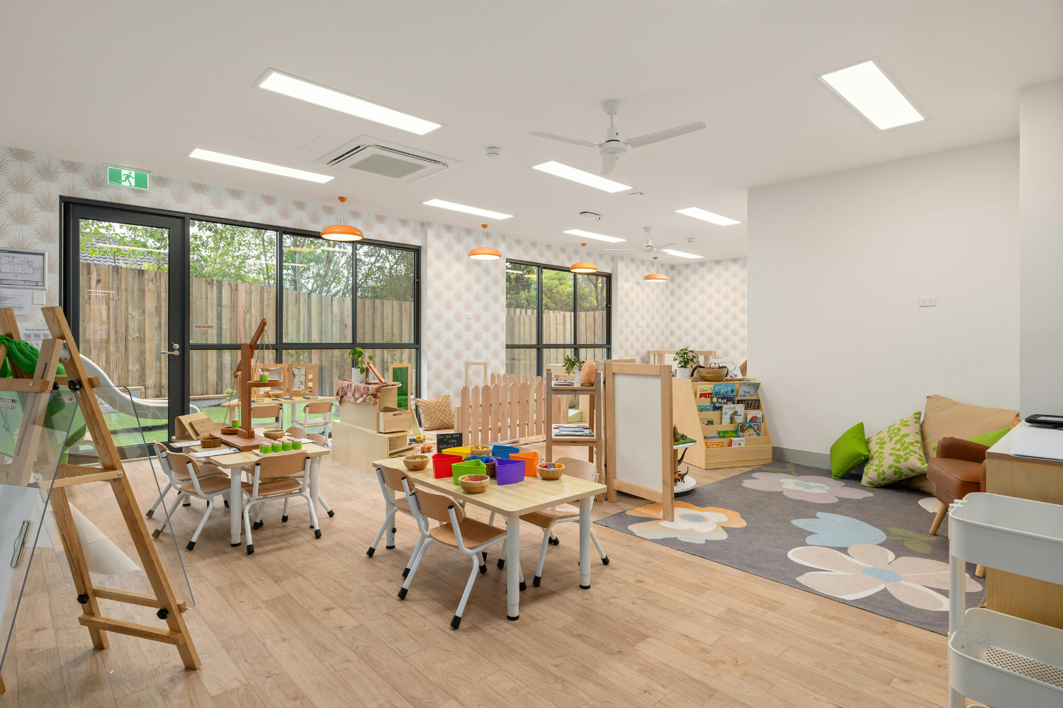 Bright children’s playroom with tables, chairs, toys, a bookshelf, and a cozy seating area with pillows and a floral rug, large windows, and a door leading outside.