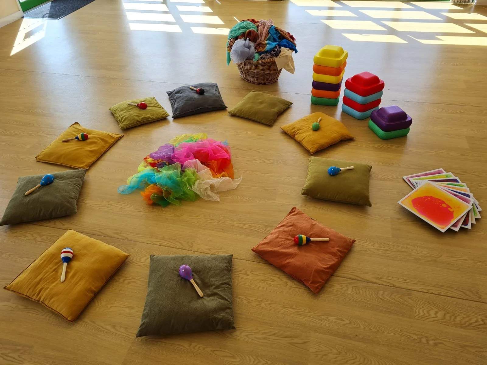 Children's circle time setup with colorful cushions, toy maracas, rainbow-colored scarves, and flashcards on a wooden floor.