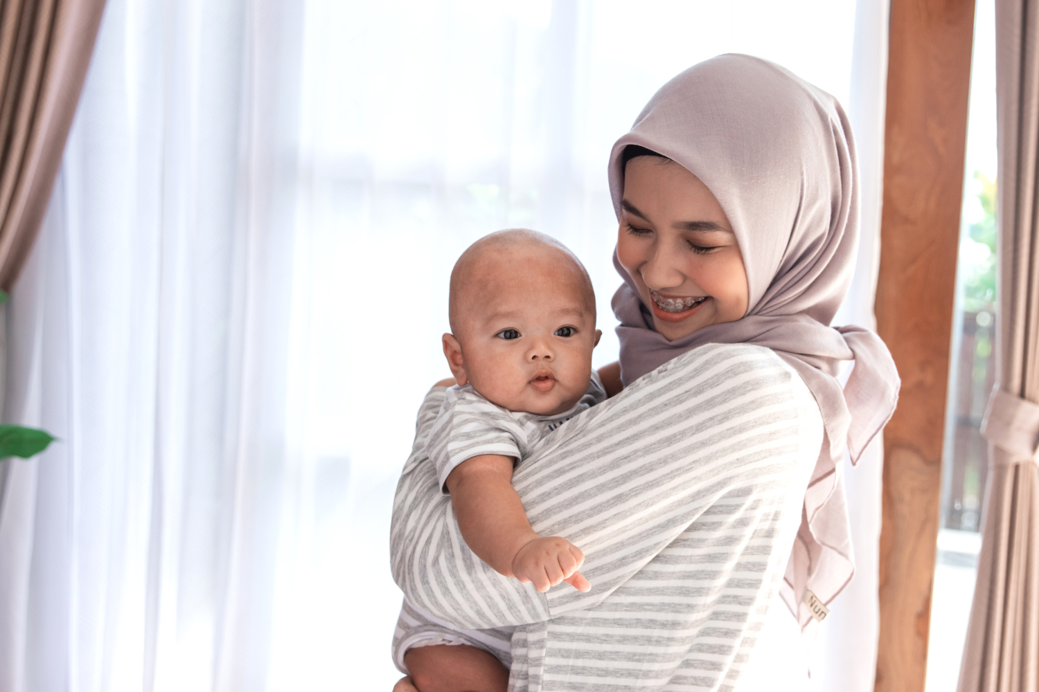 A woman wearing a beige hijab smiling and holding a baby with a bald head indoors near white curtains and wooden beams.