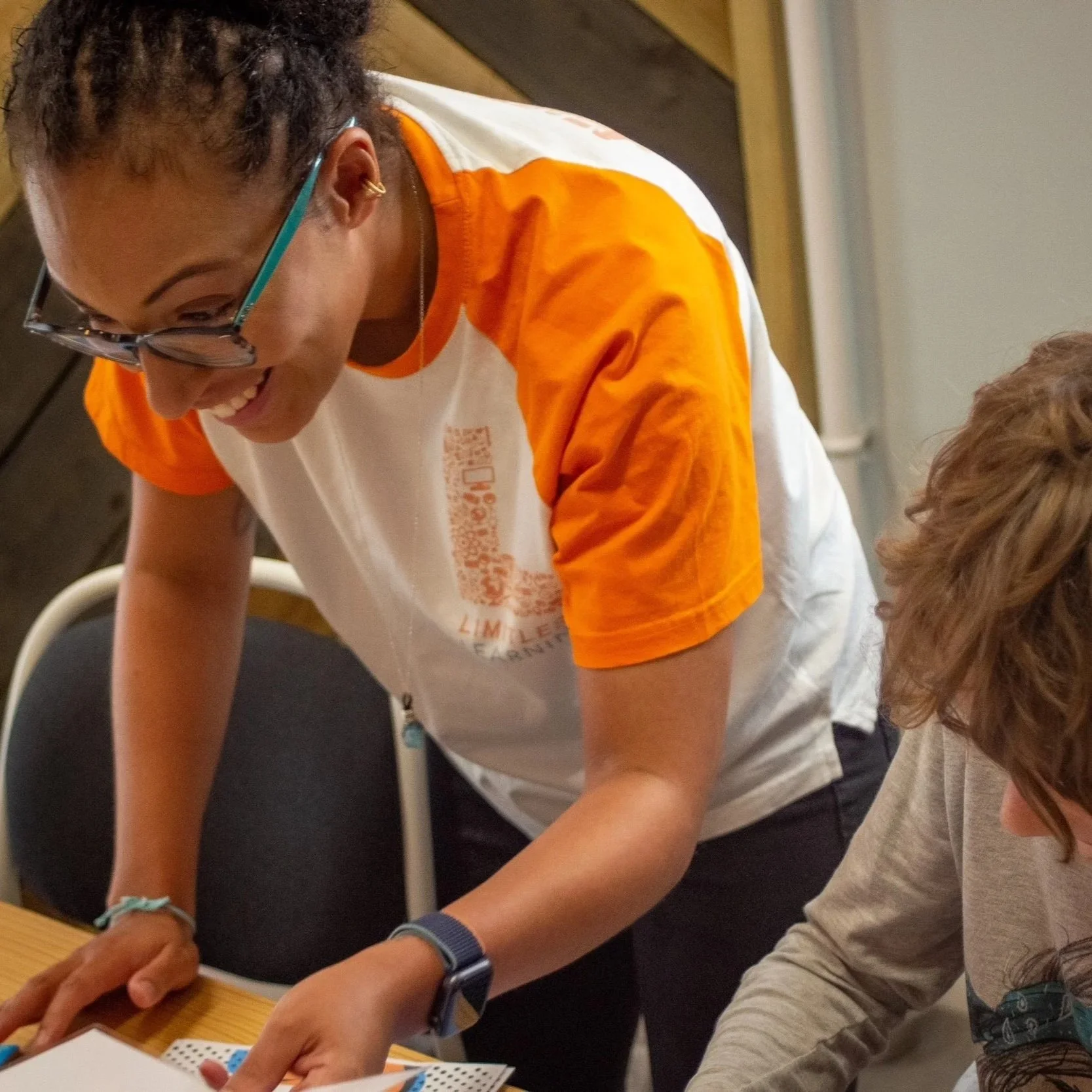 A young woman with glasses and hoop earrings, wearing an orange and white T-shirt, is leaning over a table, writing or drawing on paper, while a child with brown hair, wearing a light-colored shirt, is also focused on the activity.
