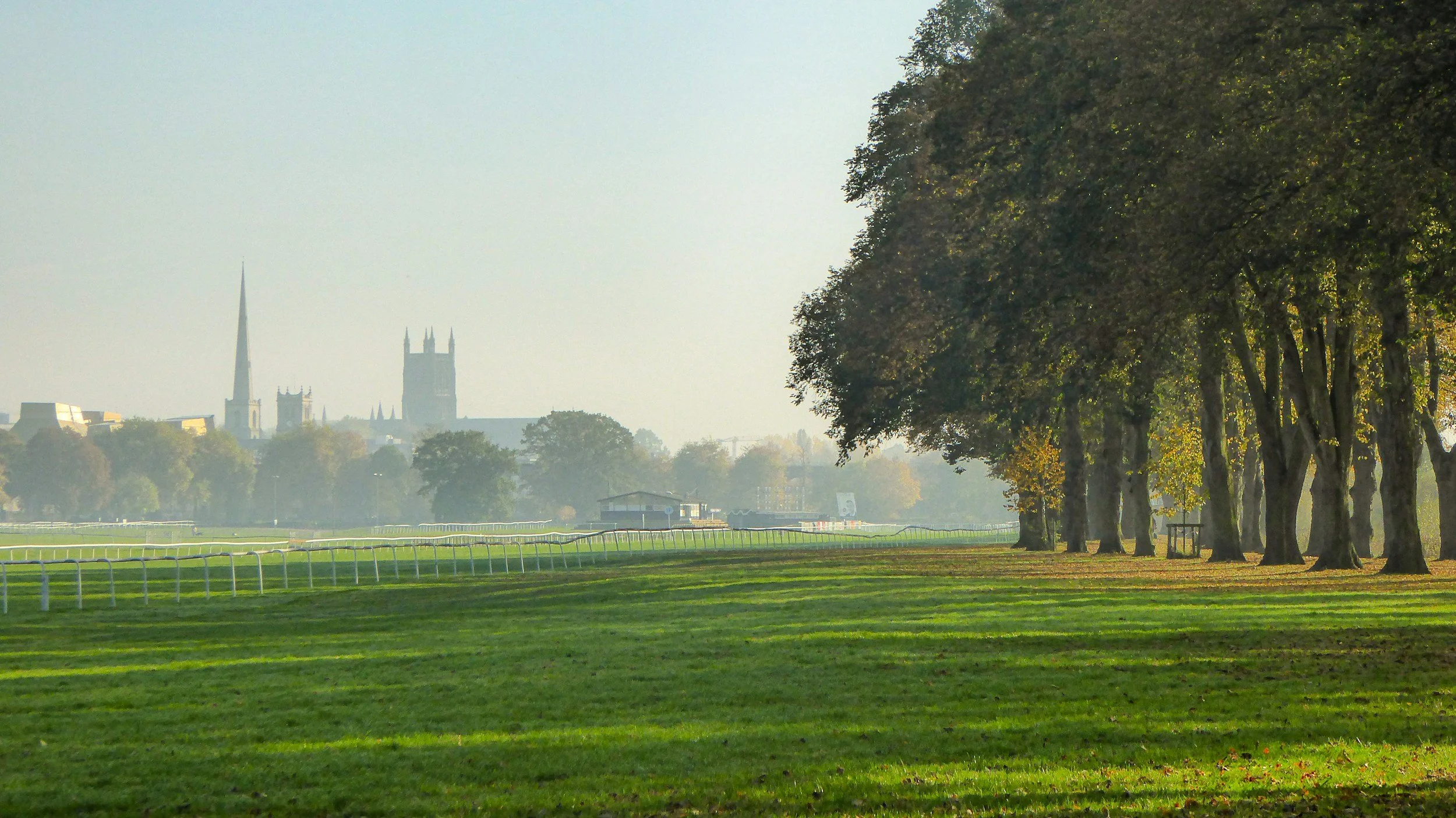 A racetrack with green grass and a row of tall trees on the right side, with a city skyline including church spires and a clock tower in the distance