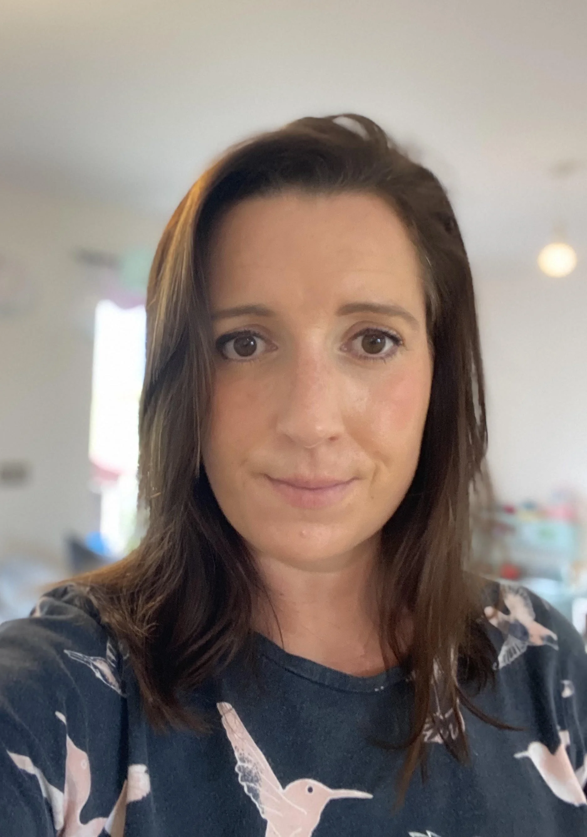 A woman with brown hair and brown eyes taking a selfie, wearing a navy blue shirt with a pink flamingo pattern in an indoor setting.