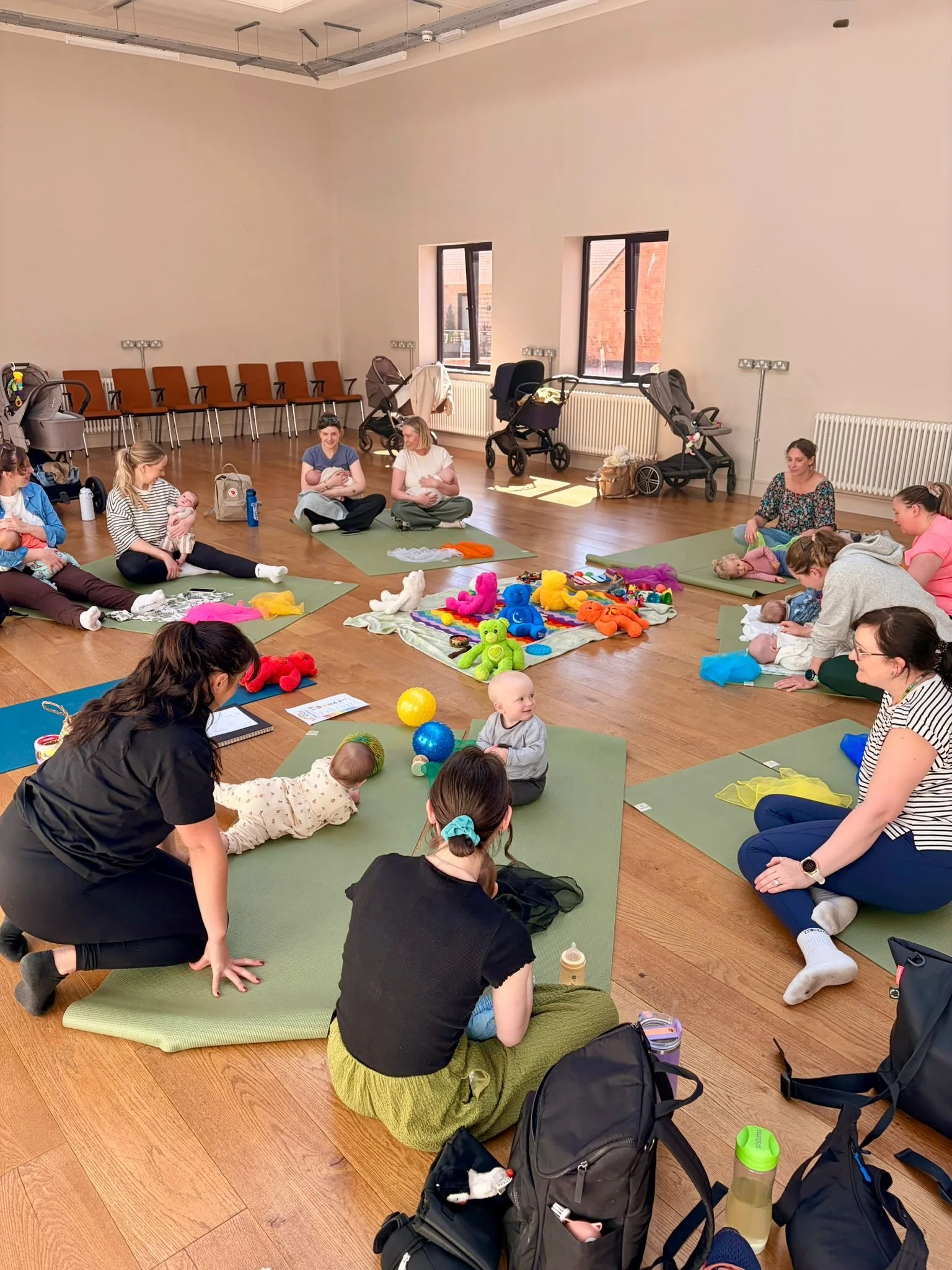 A group of women and babies participating in a baby yoga or baby massage class in a spacious room with wooden floors, large windows, and stackable chairs against the wall. The women are sitting on yoga mats, some holding or attending to their babies, surrounded by toys and baby supplies.