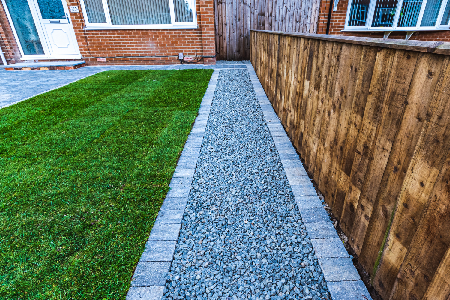 Concrete pathway bordered with bricks, leading along a wooden fence to the backyard of a brick house with a white door and windows.