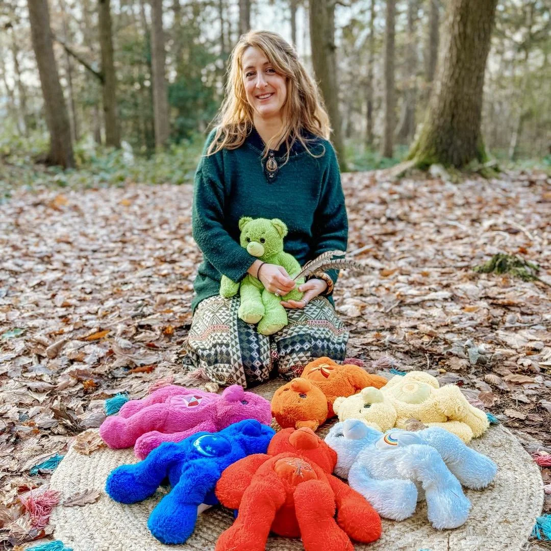Woman sitting outdoors in a forest, holding a green teddy bear and a feather, surrounded by colorful teddy bears on a mat.