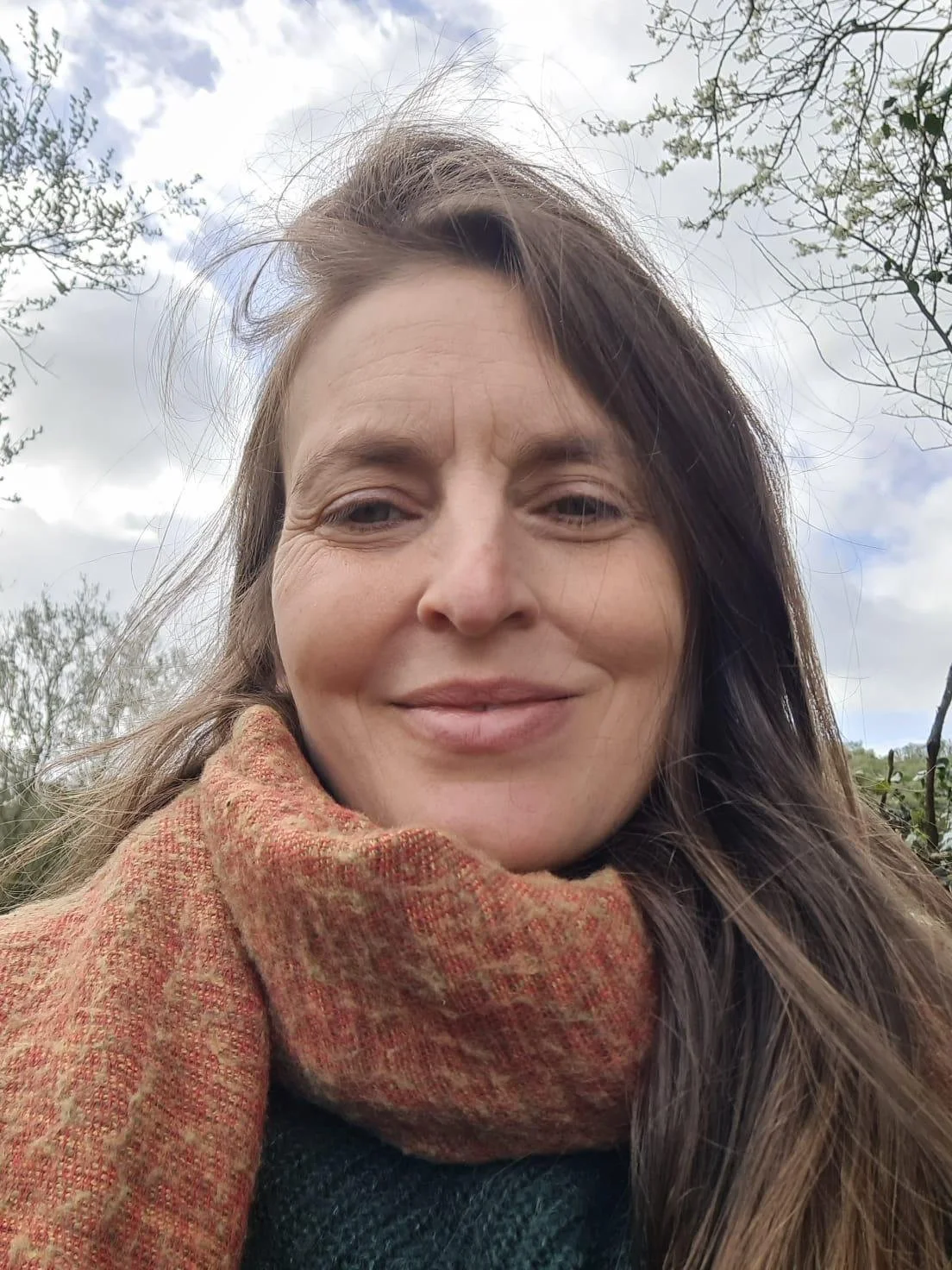 Close-up of a smiling woman with long brown hair, wearing a reddish-orange scarf, outdoors with trees and cloudy sky in the background.