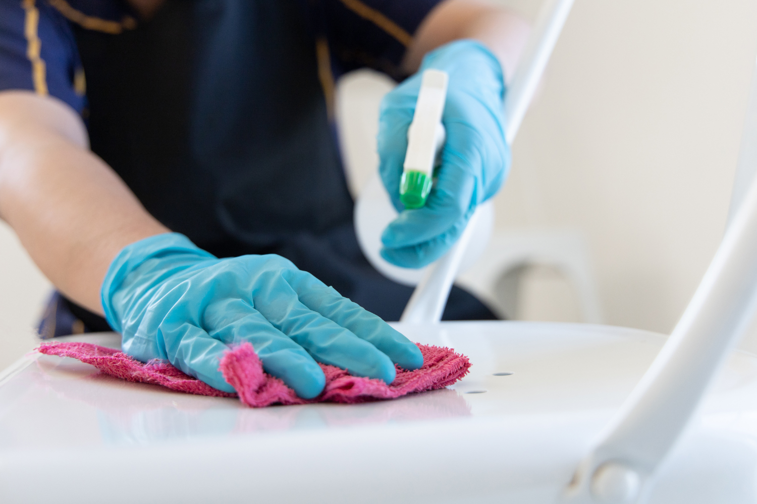 A healthcare worker wearing blue gloves is cleaning a surface with a pink cloth, holding a test tube with a green cap in one hand.